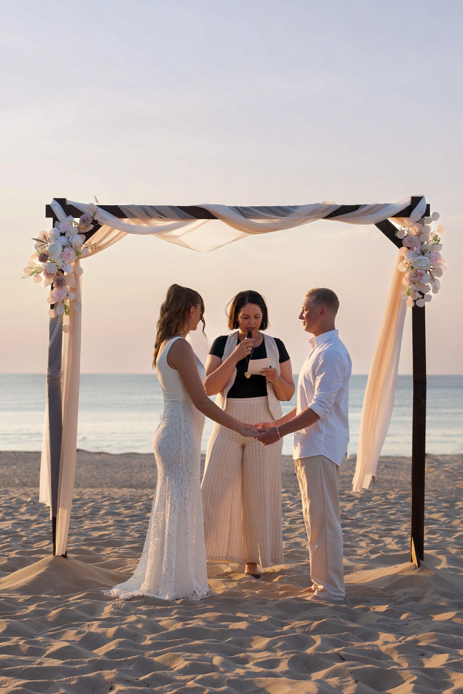 A beach wedding ceremony takes place at sunset under a white fabric canopy with flowers on a sandy shoreline.