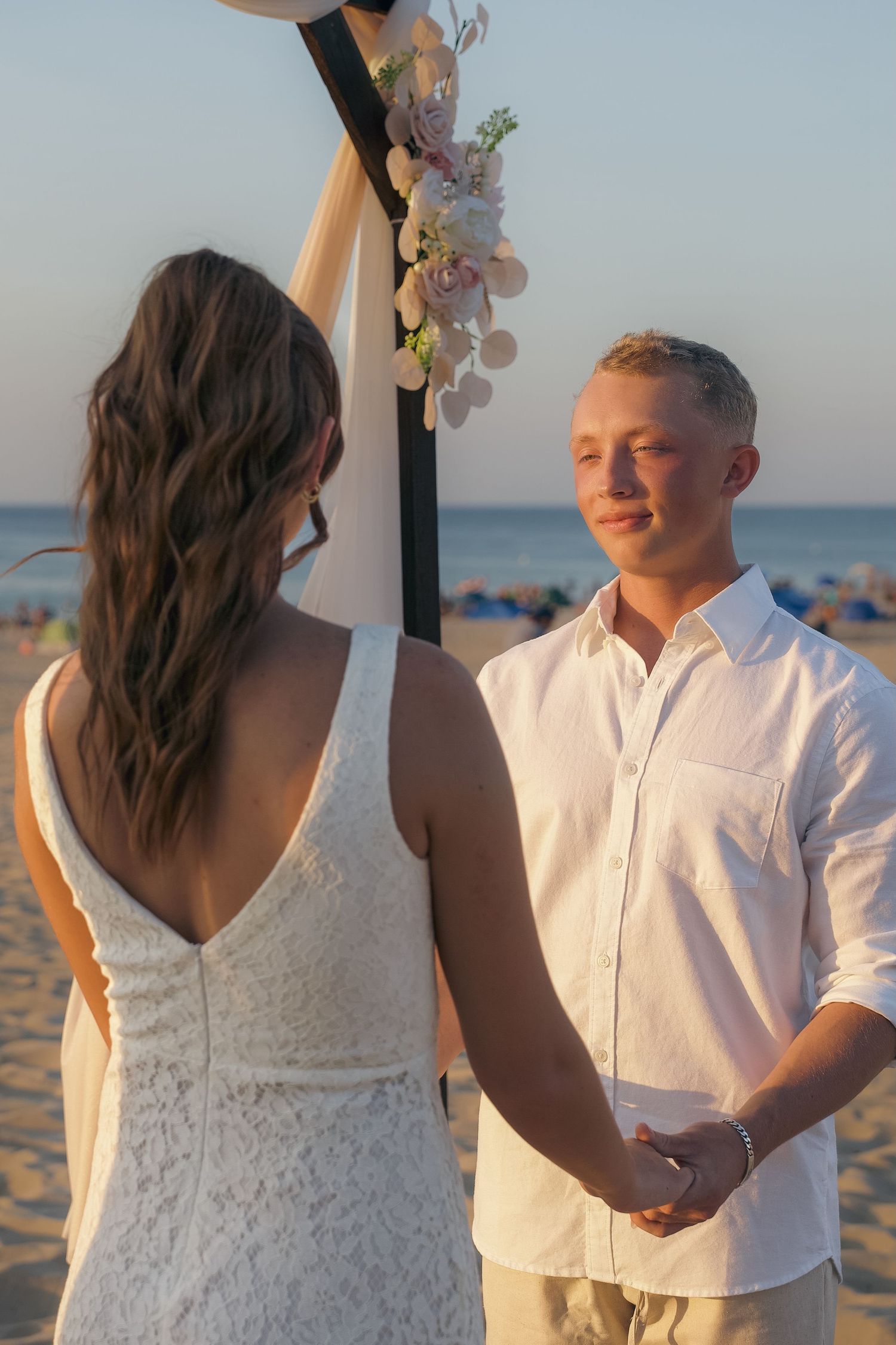 A couple holds hands during their beach wedding ceremony at sunset under a floral arch by the ocean.