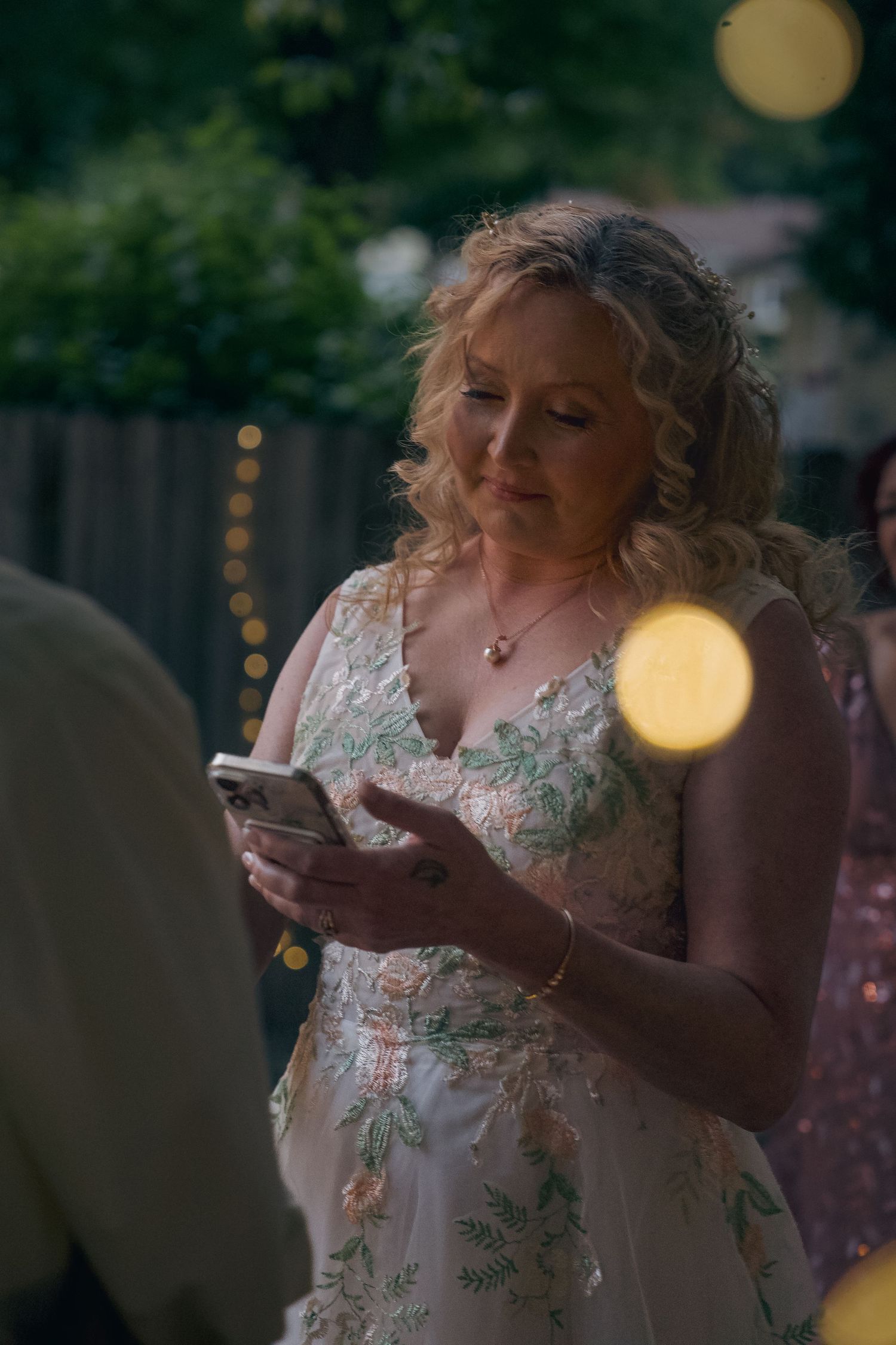 Person in a floral dress blowing bubbles outdoors at dusk with bokeh lights in the background.