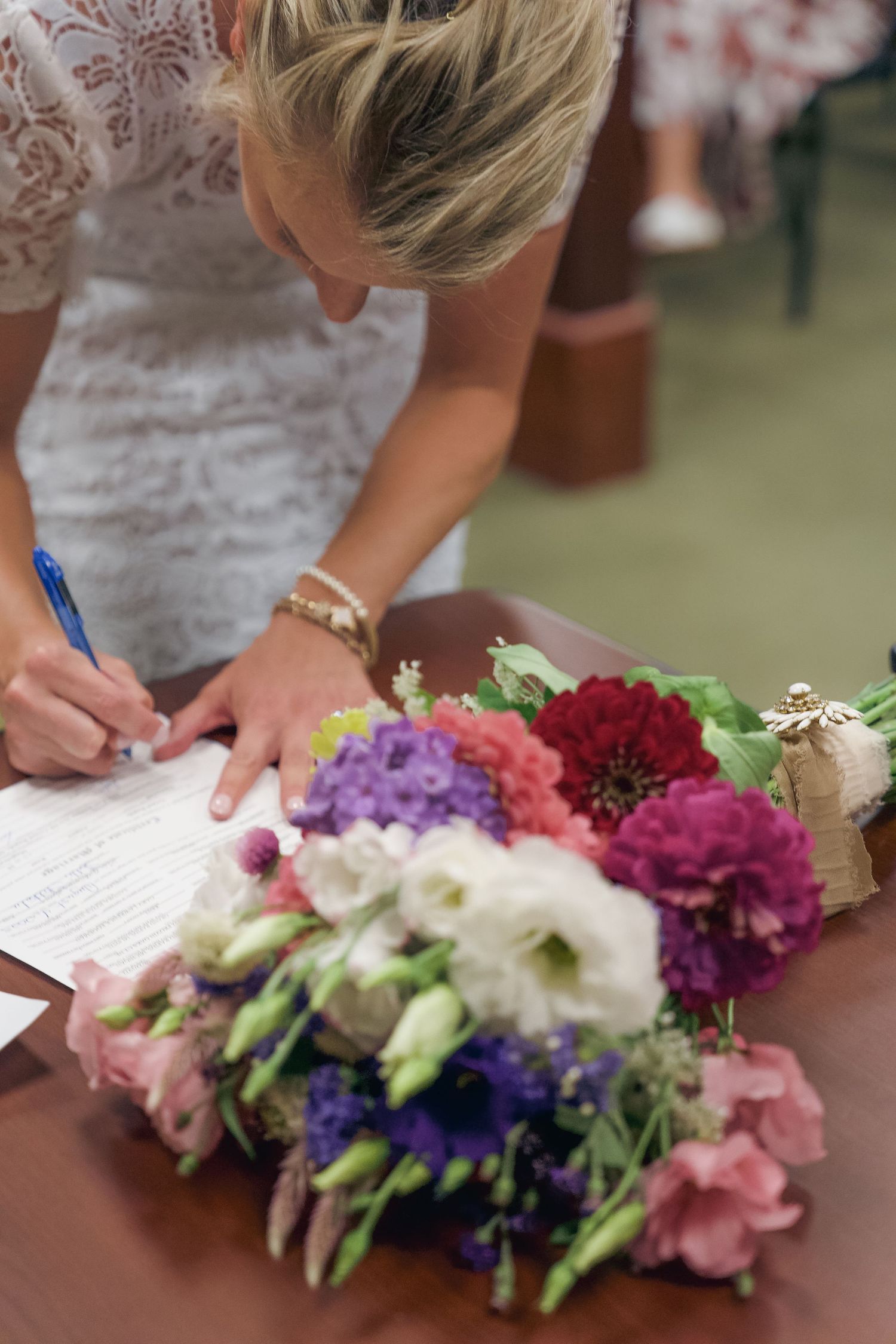 Someone in a white lace dress signs documents with colorful floral arrangements on the table during a wedding ceremony.