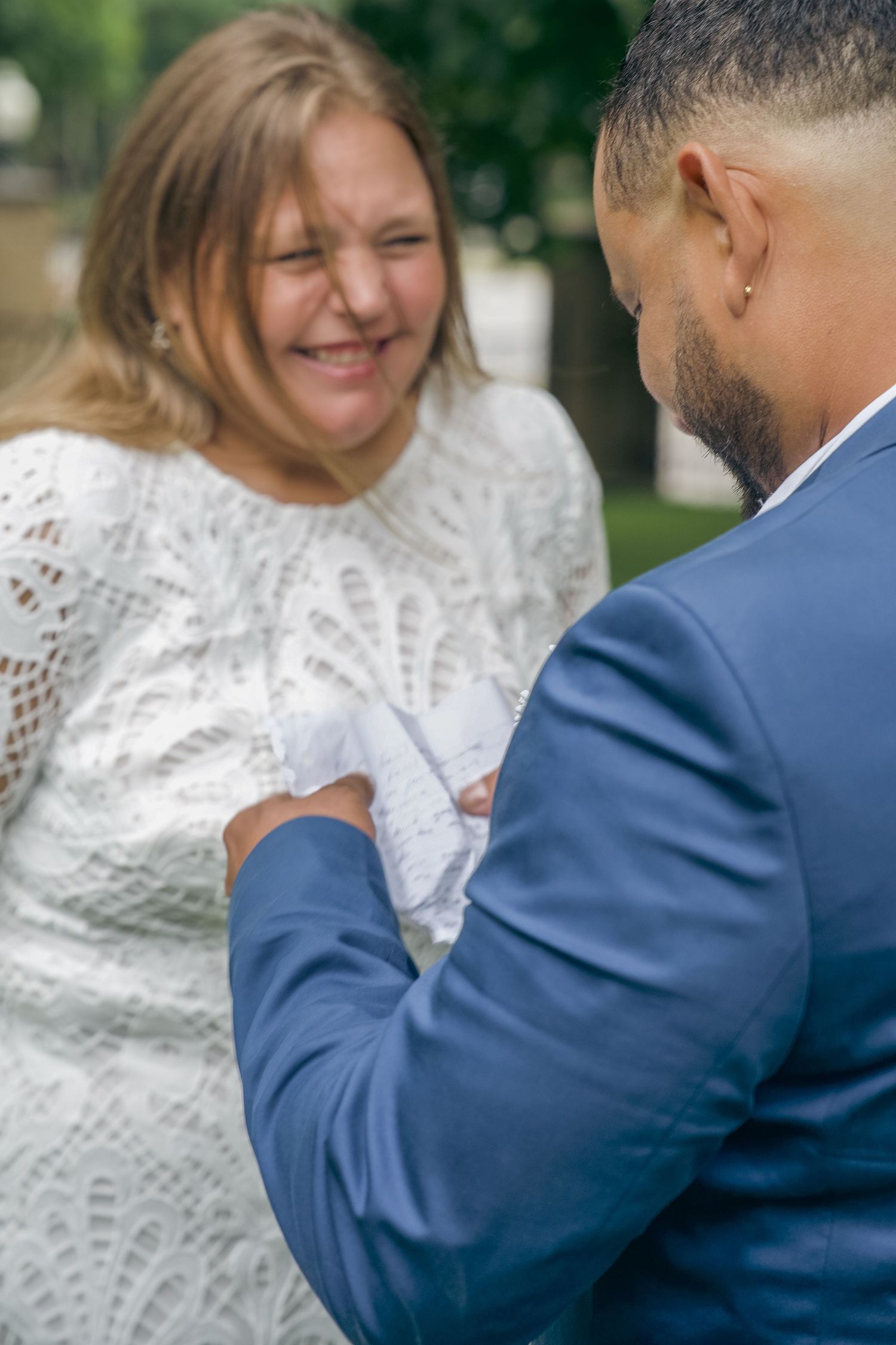 A couple in wedding attire share a tender moment outdoors, with the bride in white lace and groom in navy blue suit.