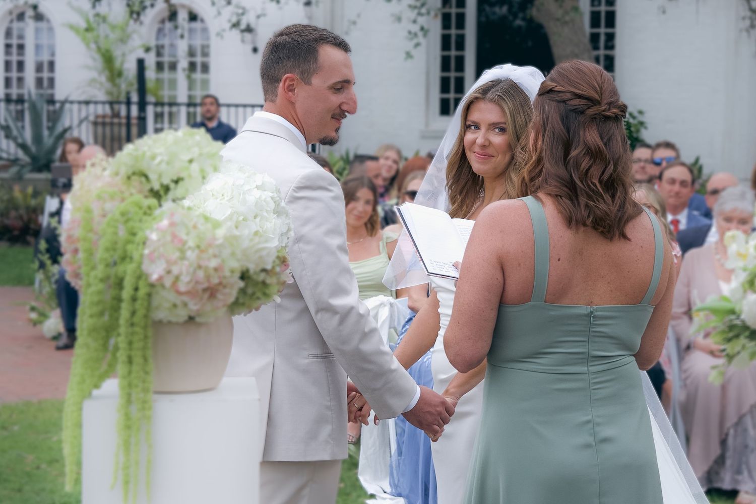 Wedding guests mingle and celebrate during an outdoor reception in a garden setting with white floral arrangements.