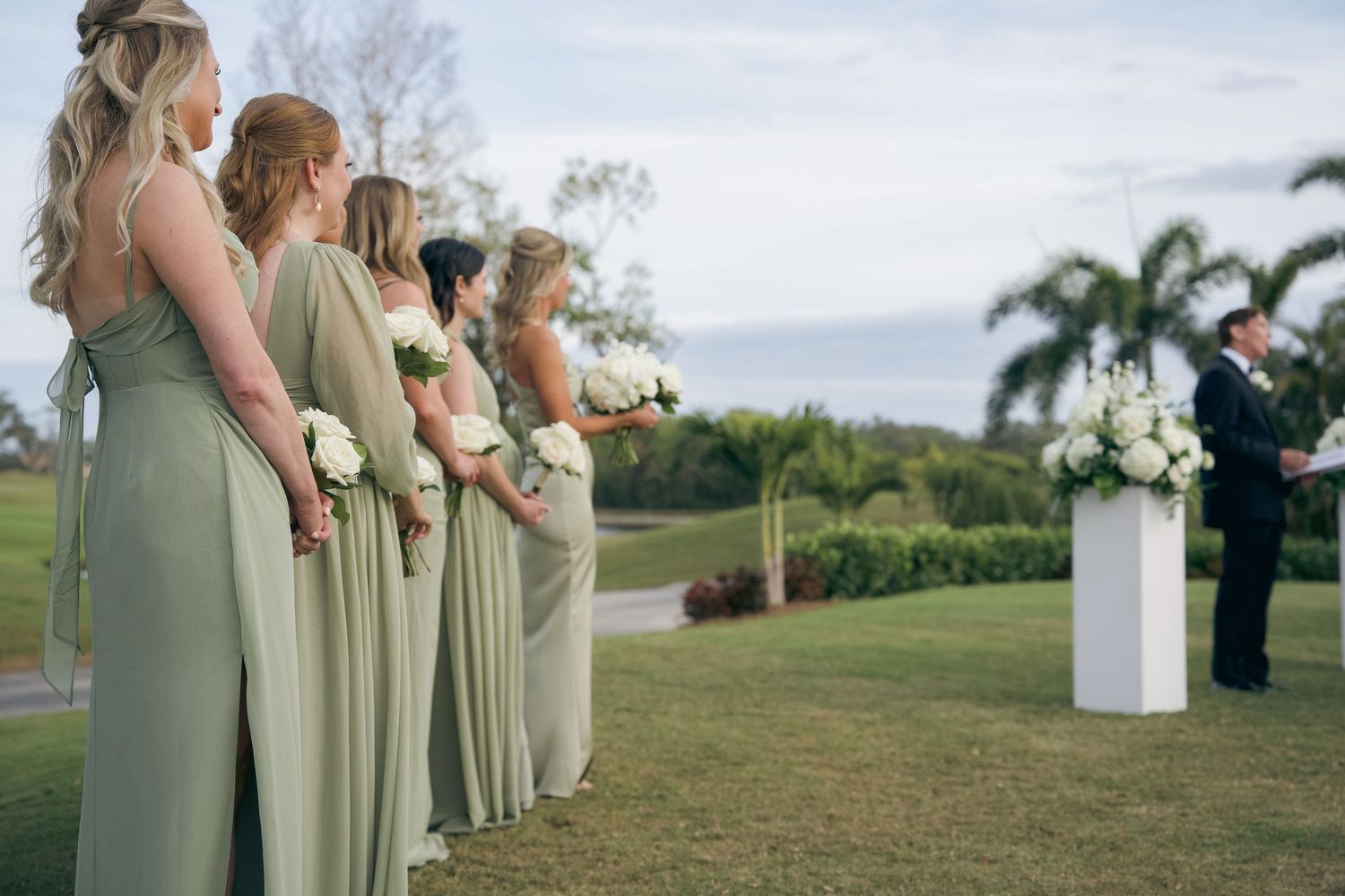 Bridesmaids in sage green dresses stand in a line at an outdoor wedding ceremony with a scenic landscape view.