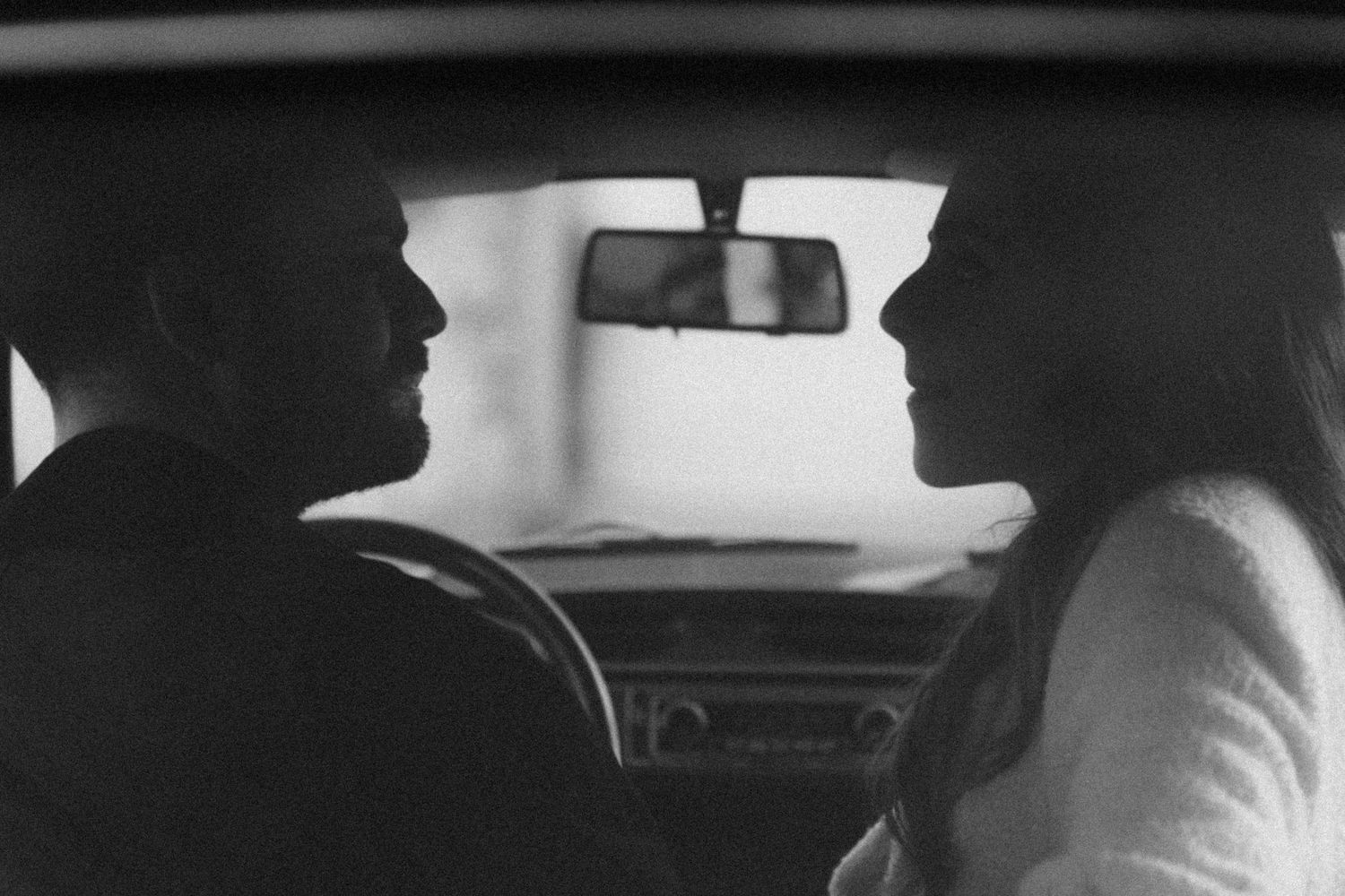 Monochrome image capturing a side view of two silhouetted figures seated in a vehicle interior.
