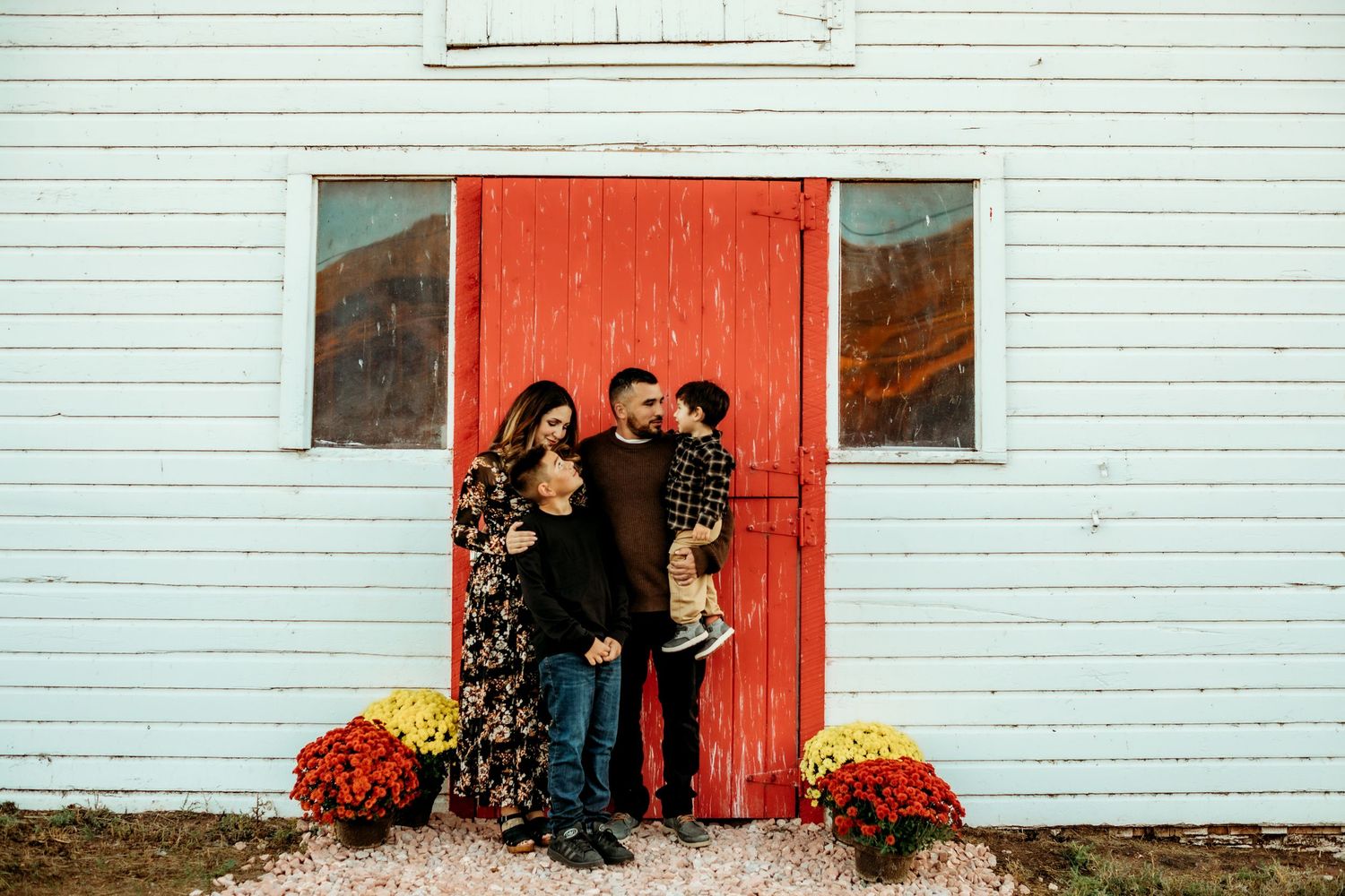 A couple stands in front of a bright red door on a white building with orange mums decorating the entrance.