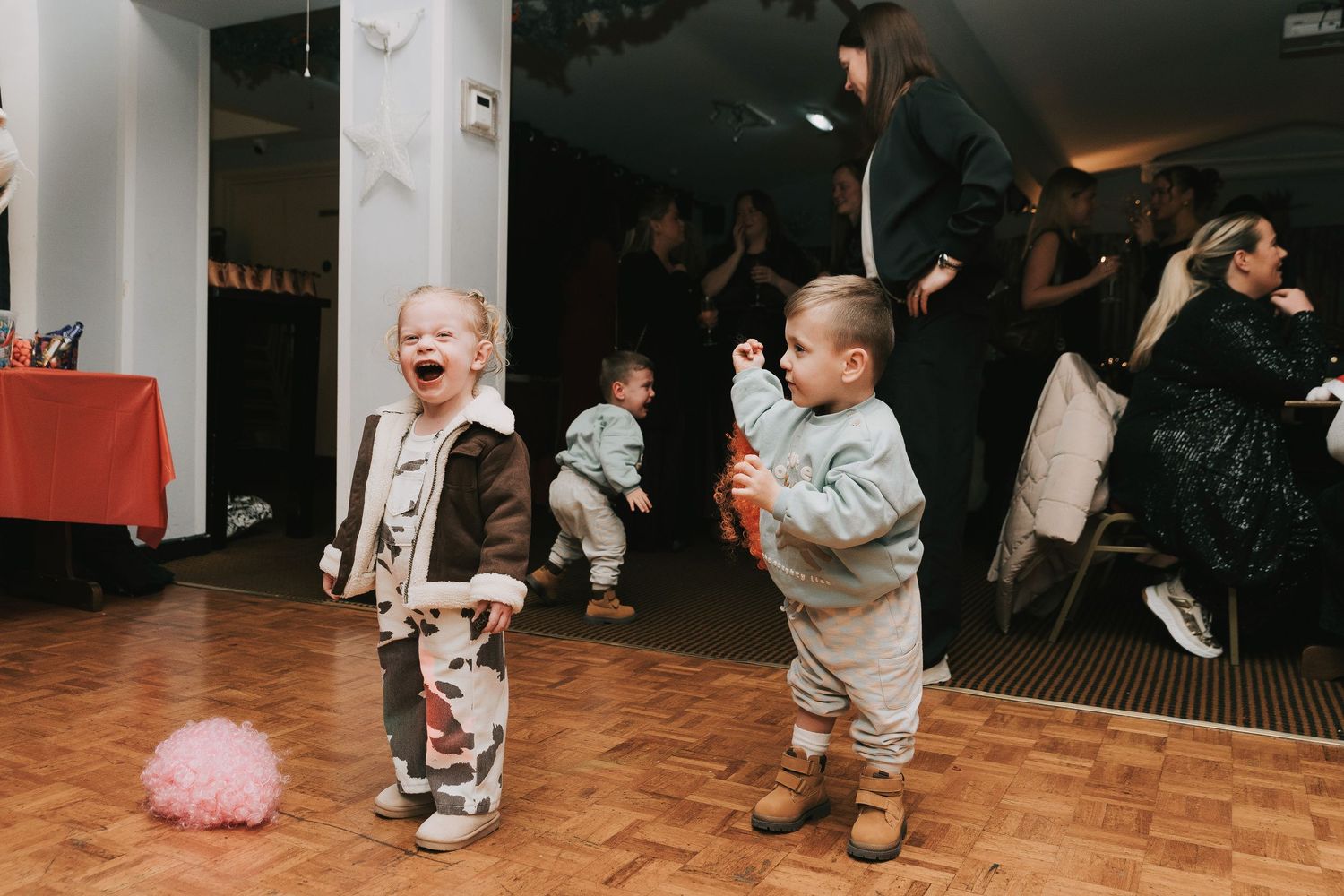 Two toddlers dancing on a wooden floor at a party with adults in the background.