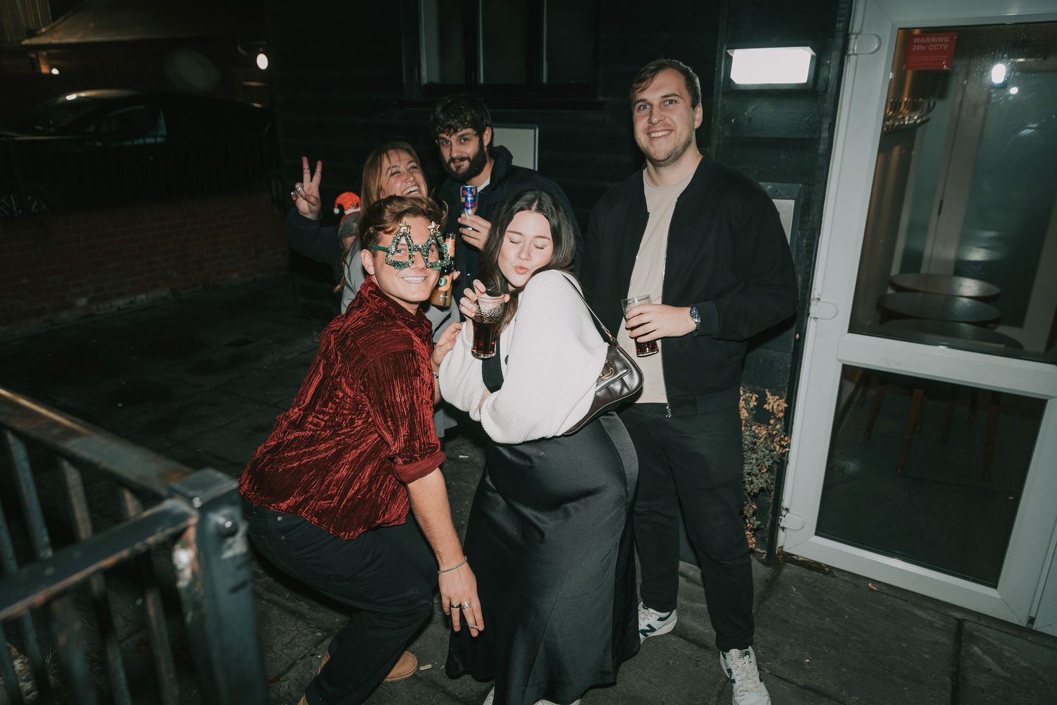 A group of friends pose together at night outside a building entrance, with one wearing a red velvet jacket.