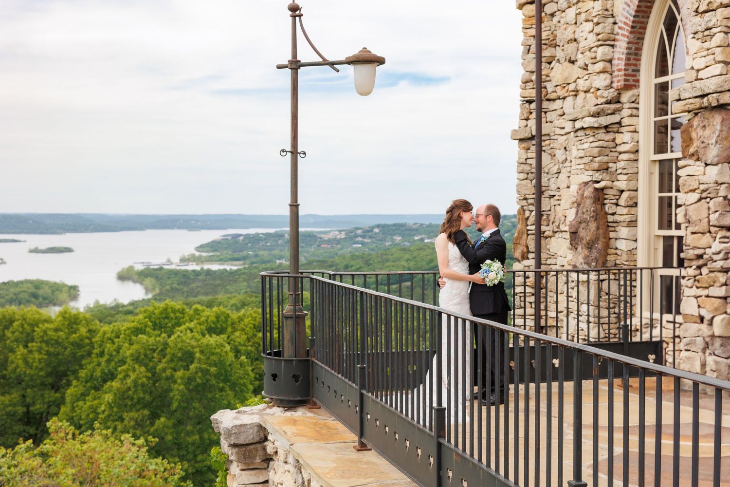 A romantic couple shares a tender moment on a scenic overlook balcony with mountains and water views in the distance.