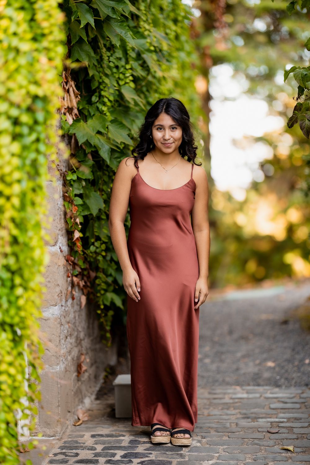 A person in a rust-colored dress poses on stone steps surrounded by lush green ivy and foliage.