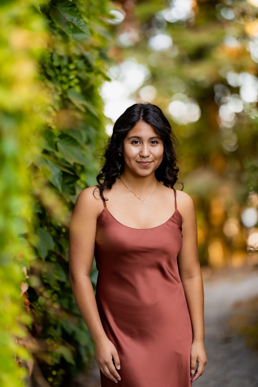 Woman in burgundy dress poses outdoors with sunlit foliage creating a warm autumn atmosphere.