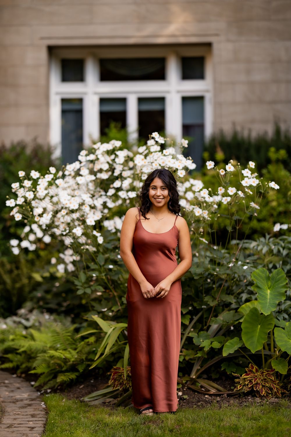 A person in a burgundy dress poses by white flowers and ferns in a garden setting.