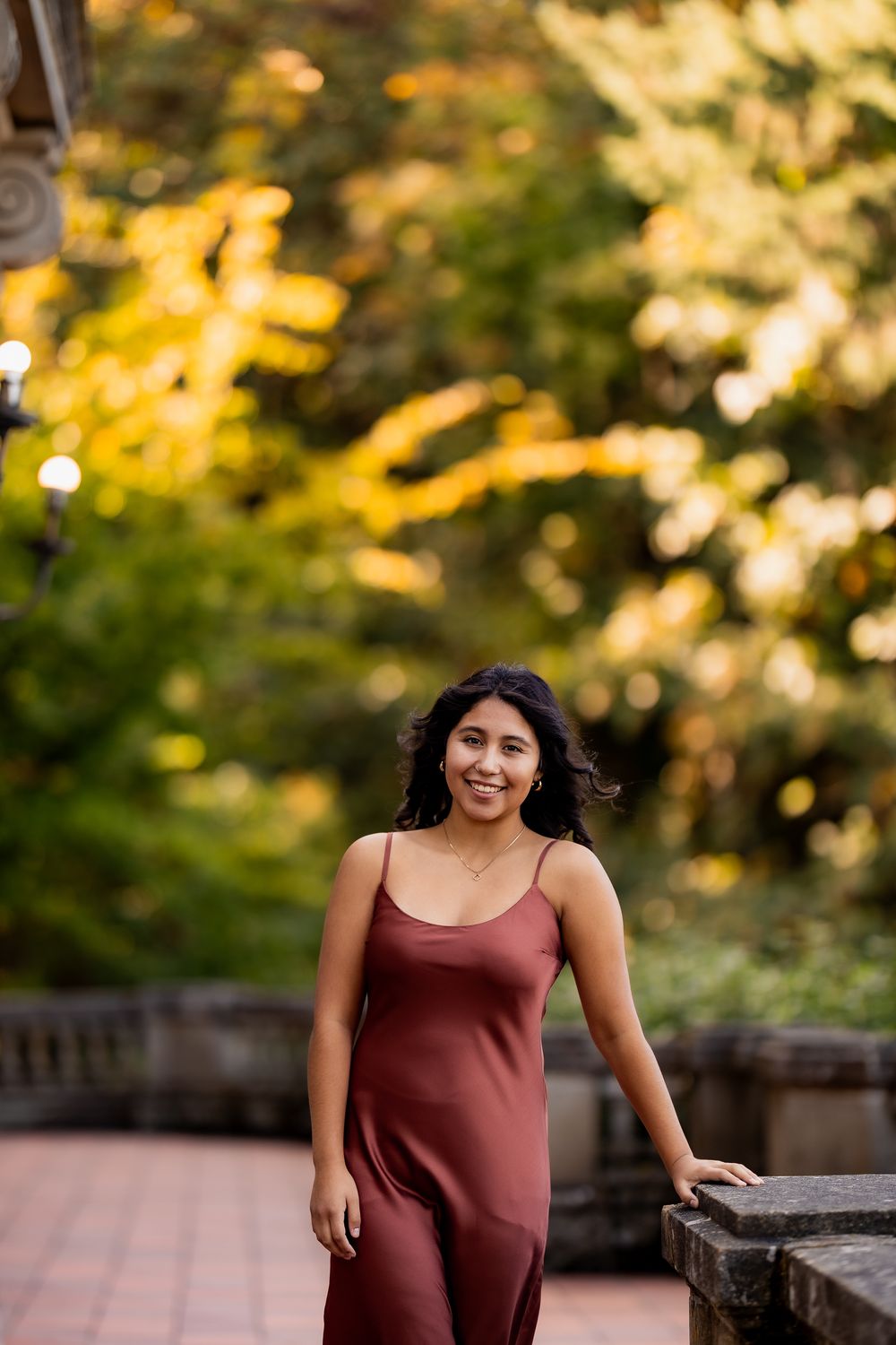 Woman in maroon dress stands near wooden railing with golden sunlight filtering through trees in background.