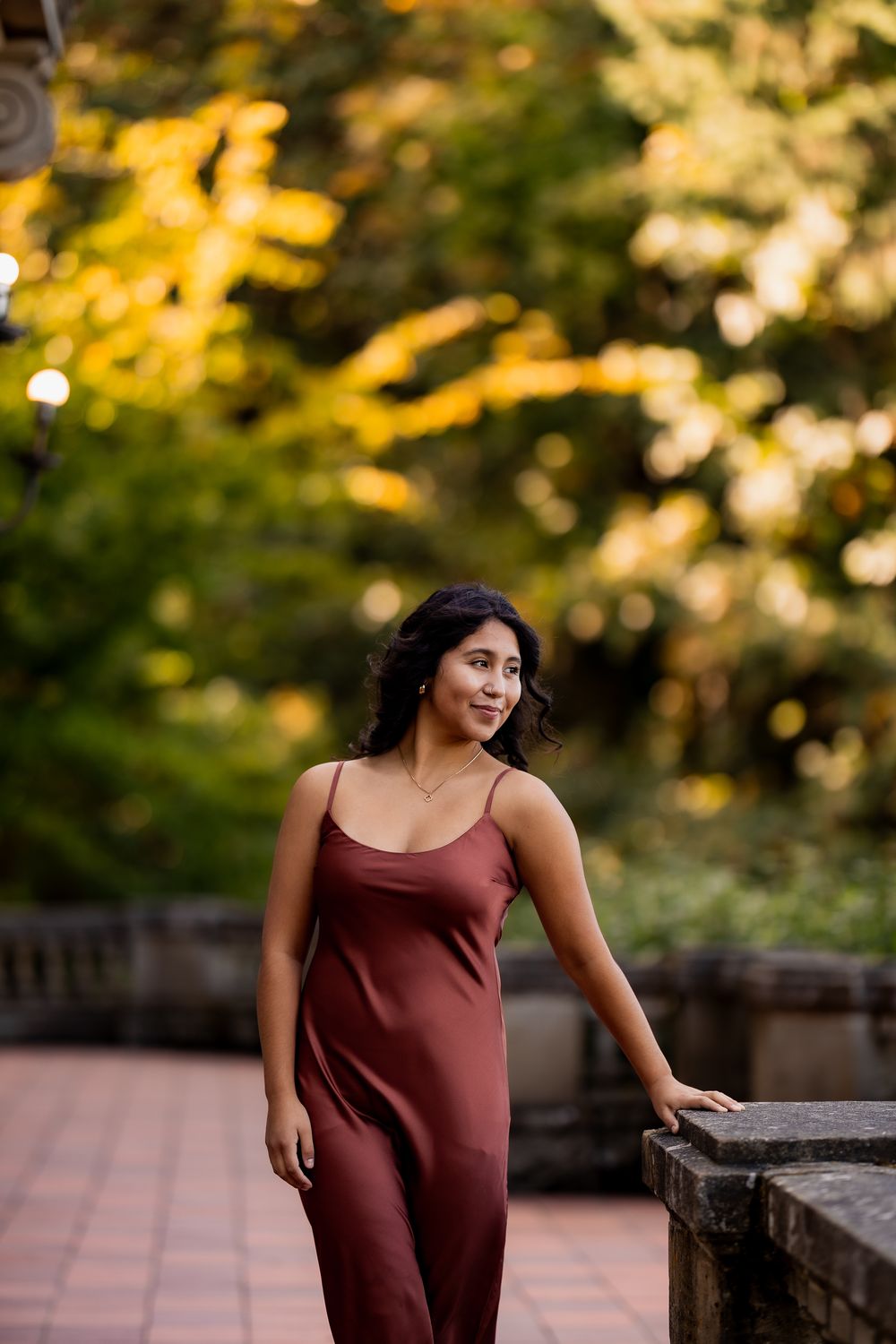A person in a dark red dress poses against autumn foliage during golden hour.
