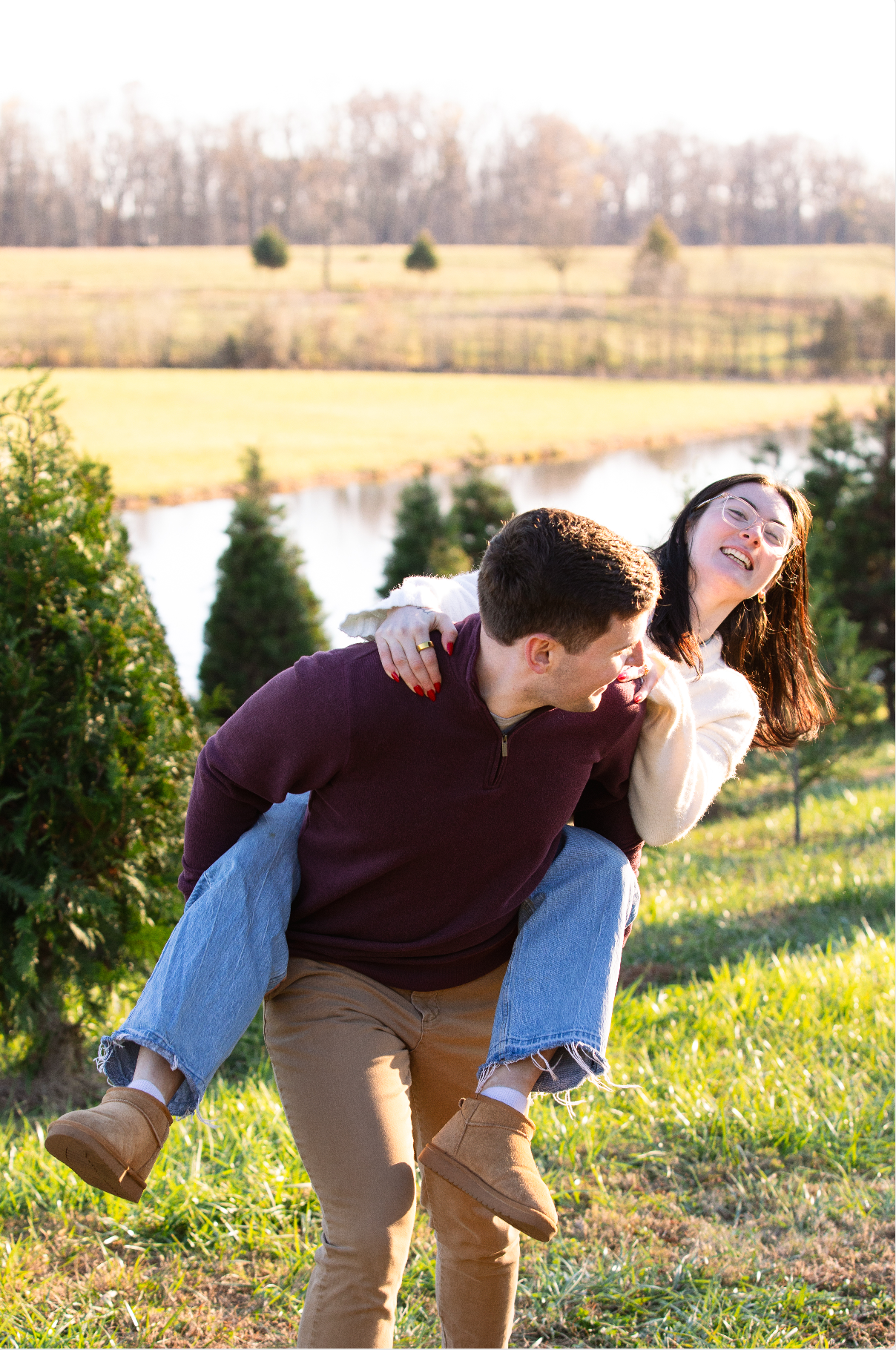 A couple playfully dips and poses together in a scenic field with a lake and trees at sunset after a proposal