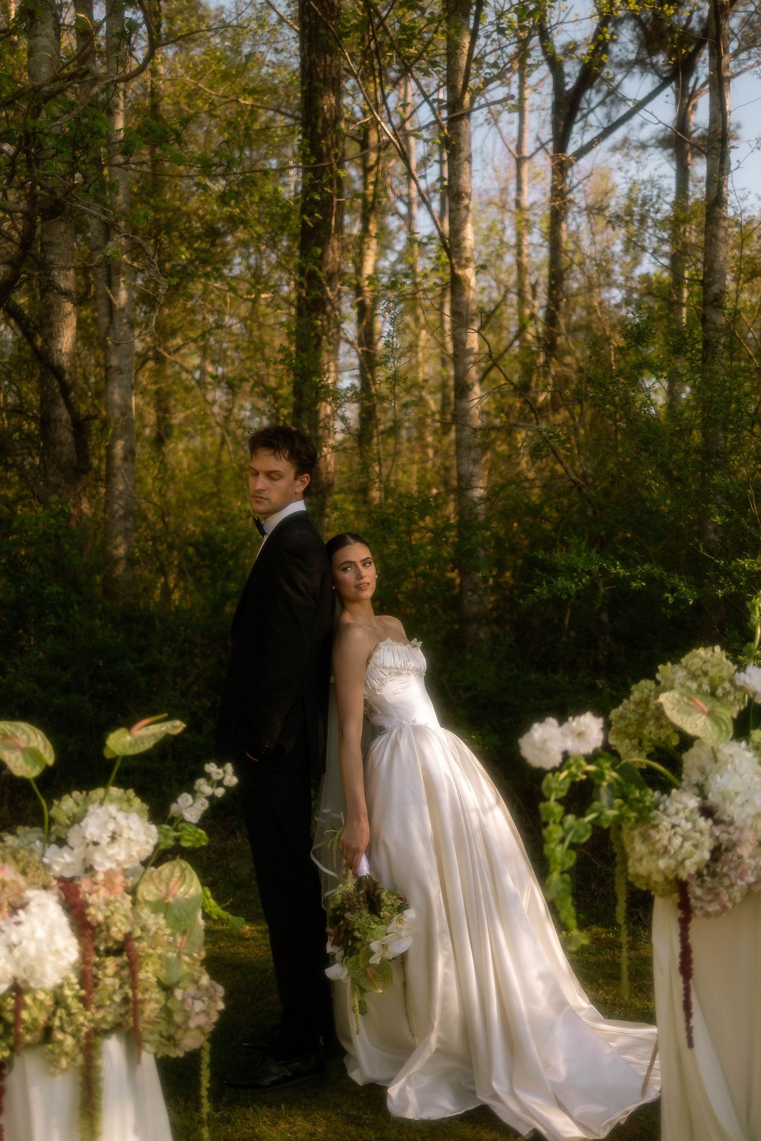 A bride and groom pose back-to-back in a lush forest wedding setting surrounded by elegant floral arrangements.