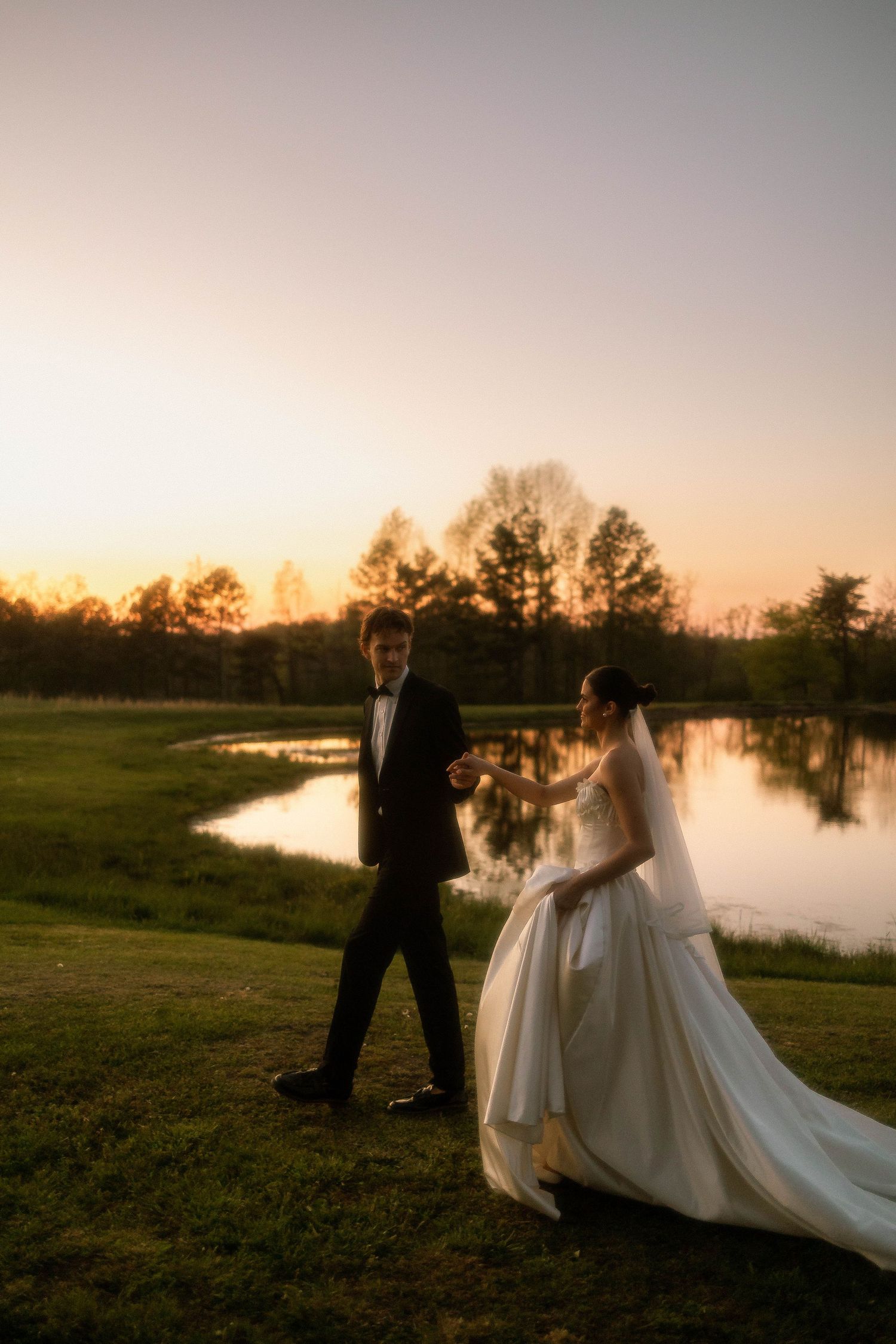 Bride and groom hold hands walking by a pond at golden hour sunset, surrounded by lush green trees.