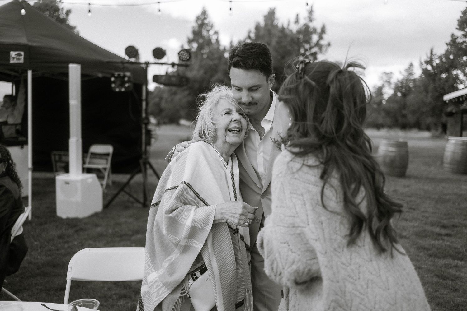 Bride and groom share a tender moment kissing an elderly woman at an outdoor wedding reception.