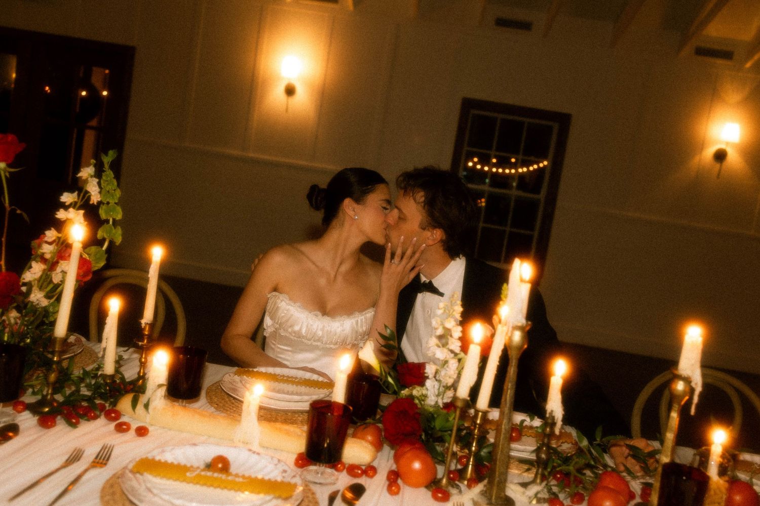 Bride and groom share a romantic candlelit kiss at their wedding reception dinner table adorned with red roses.