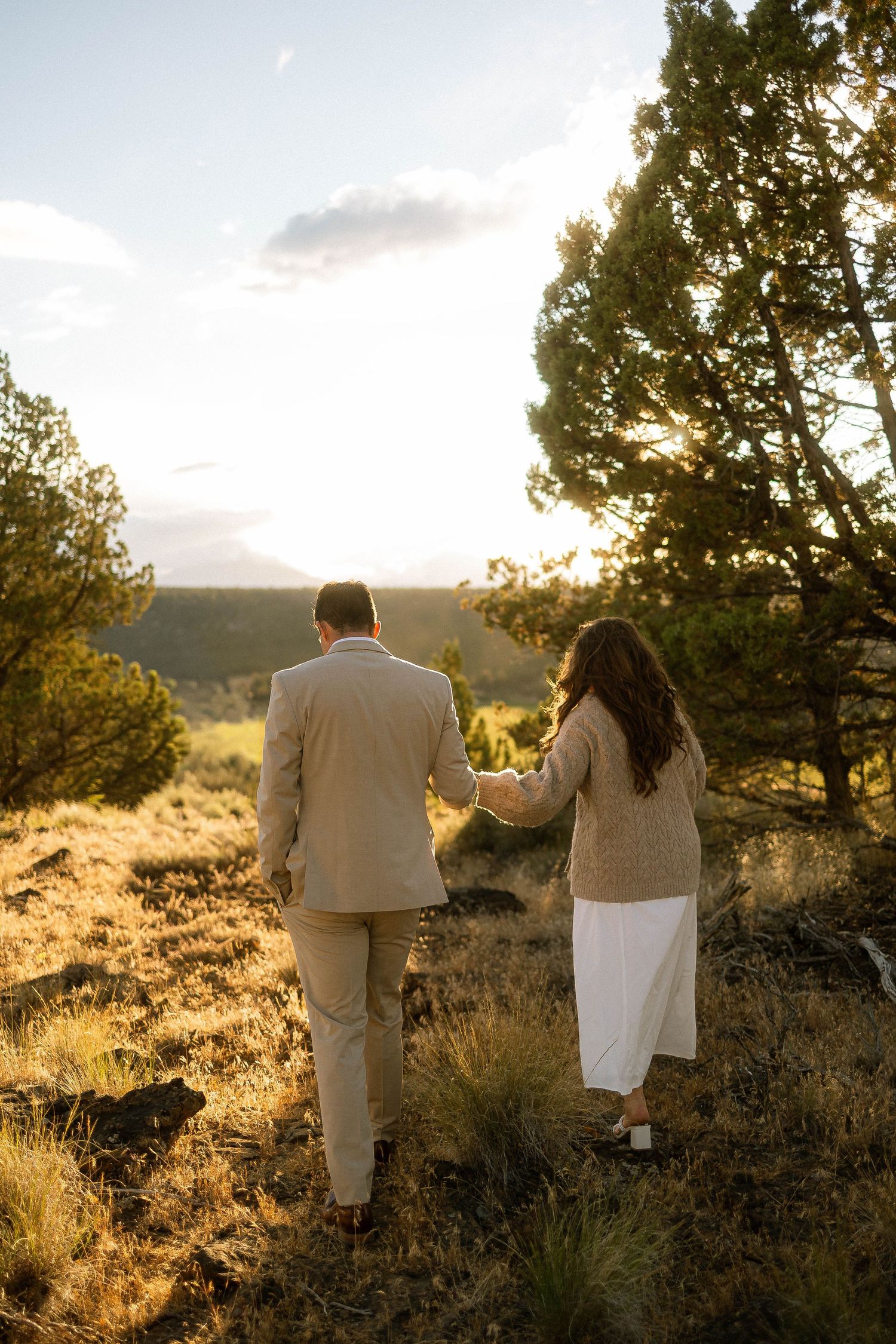 A couple holds hands walking through a sunlit forest meadow, dressed in elegant neutral tones at golden hour.