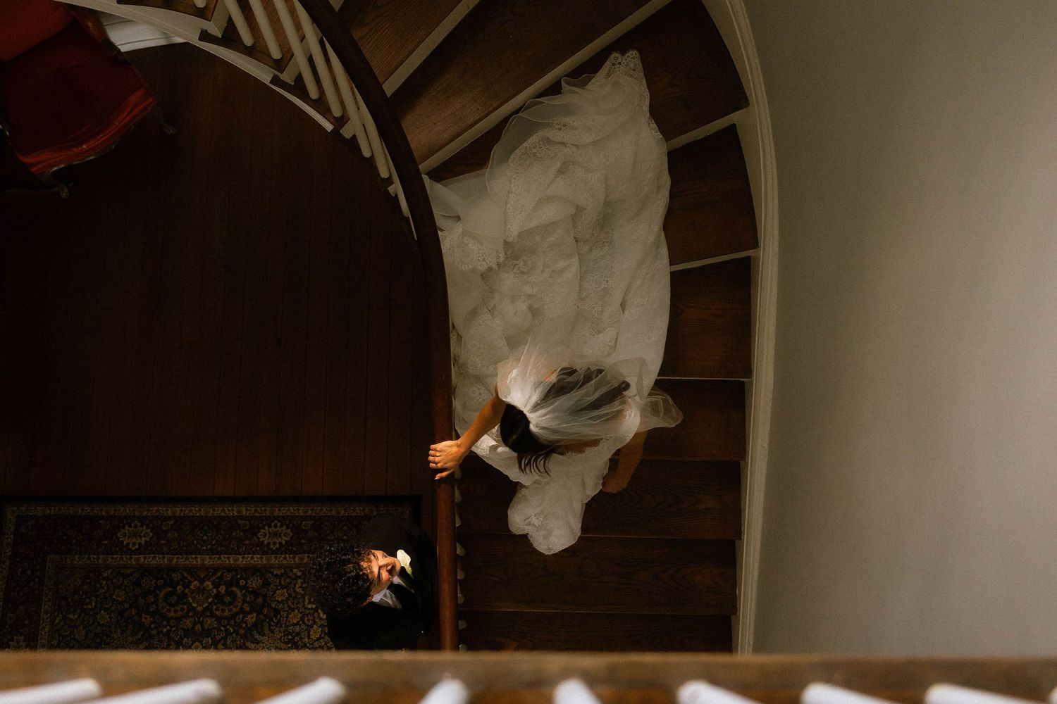 Aerial view of a bride in a white wedding gown descending a wooden spiral staircase, her dress flowing elegantly.