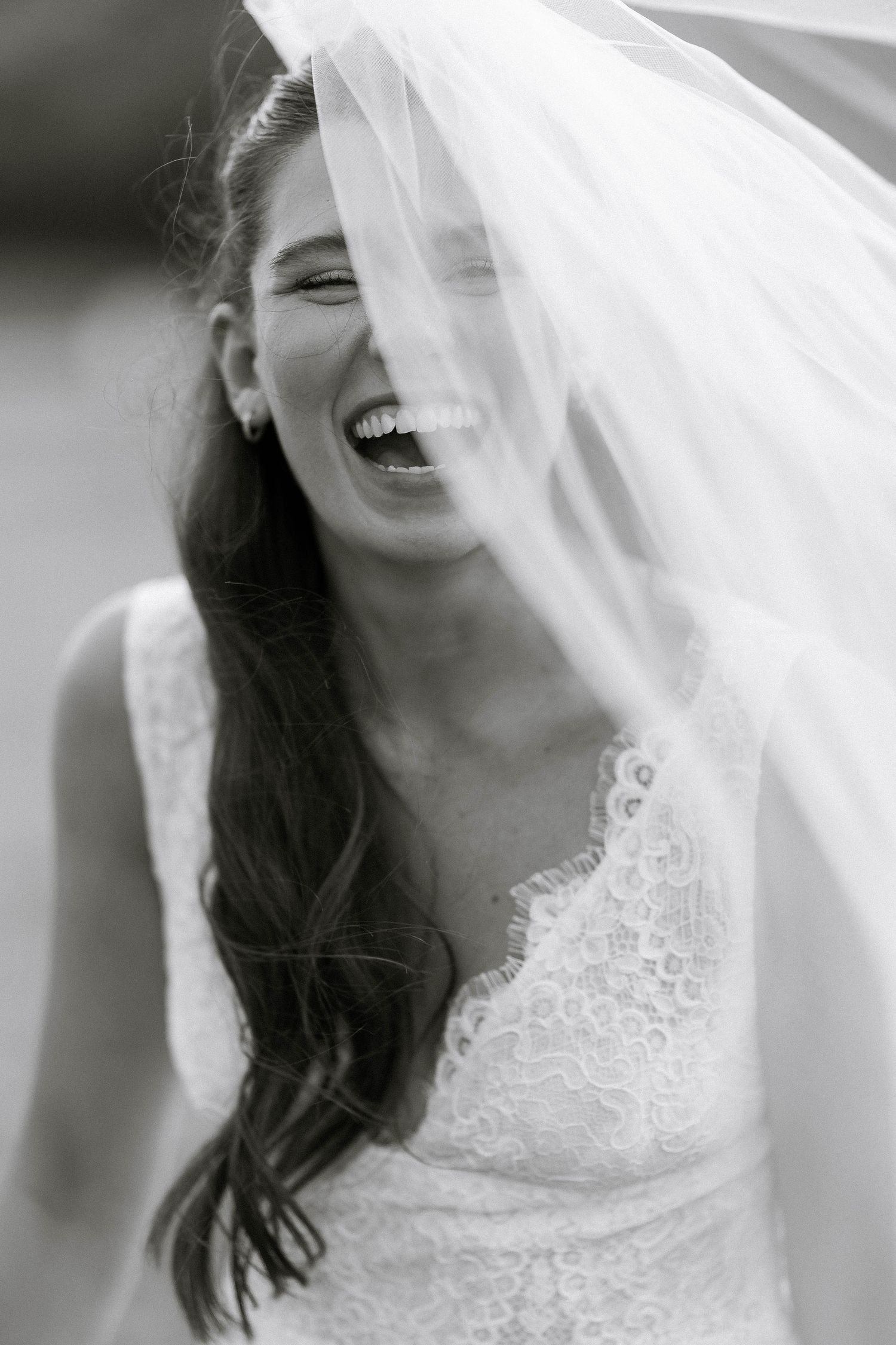 Black and white photo of a laughing bride in lace dress with veil blowing across her face on her wedding day.