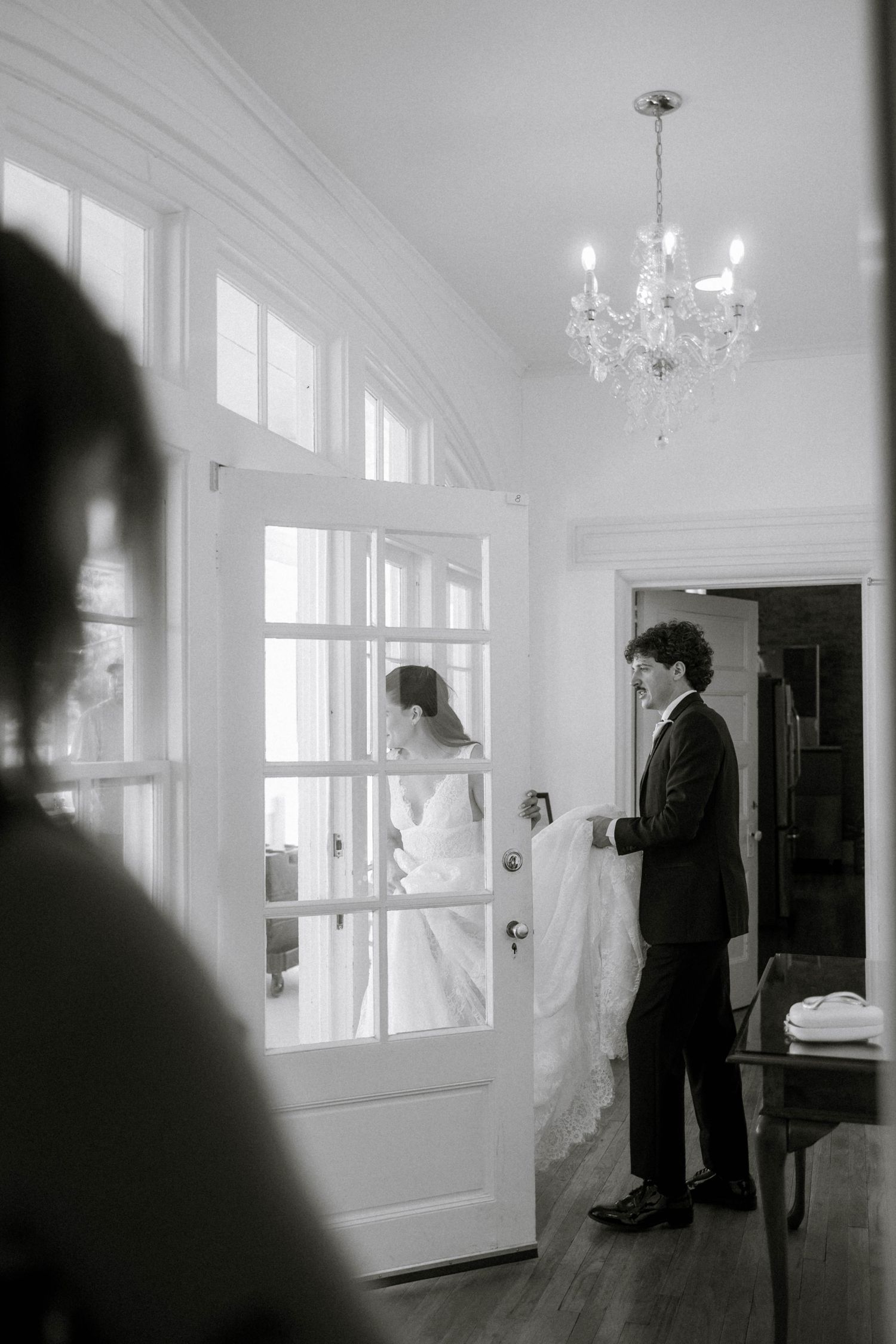 Black and white photo of a groom holding a bride's dress train near French doors, with a chandelier above.
