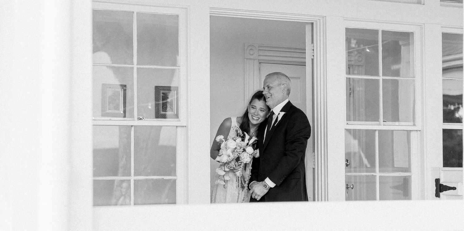 Black and white photo of a bride and groom smiling together at an elegant white French door entrance.