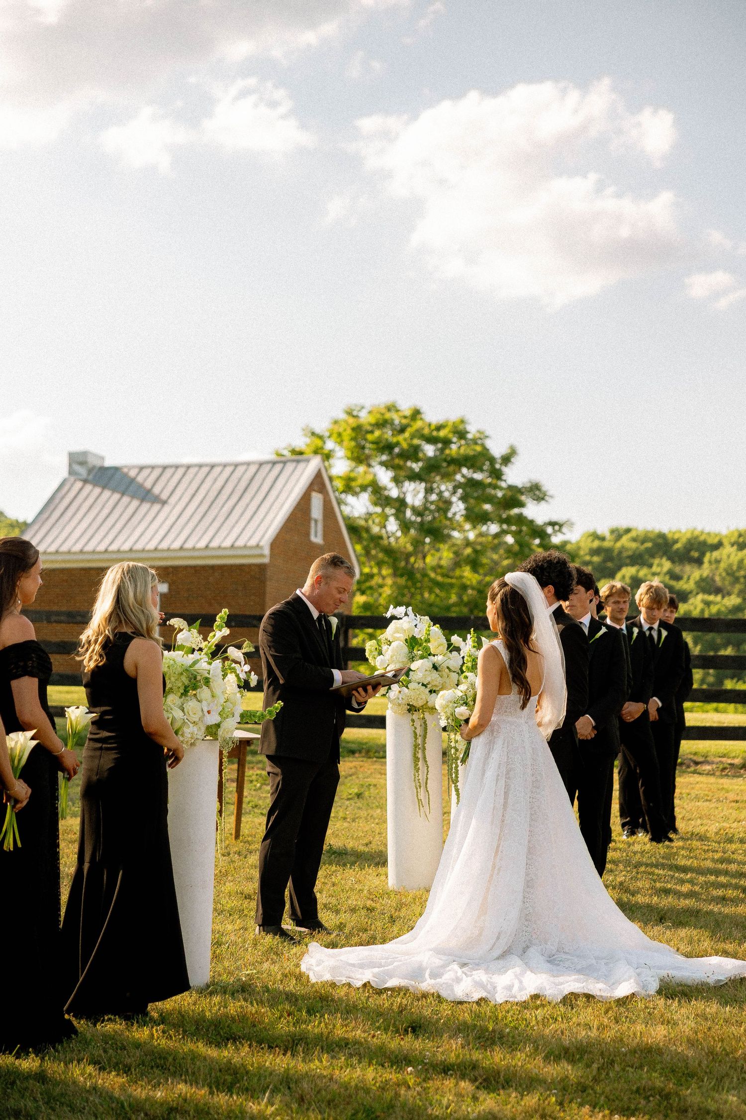 Outdoor wedding ceremony on green lawn with bride in white gown, groom, and wedding party in black attire beside floral arrangements.