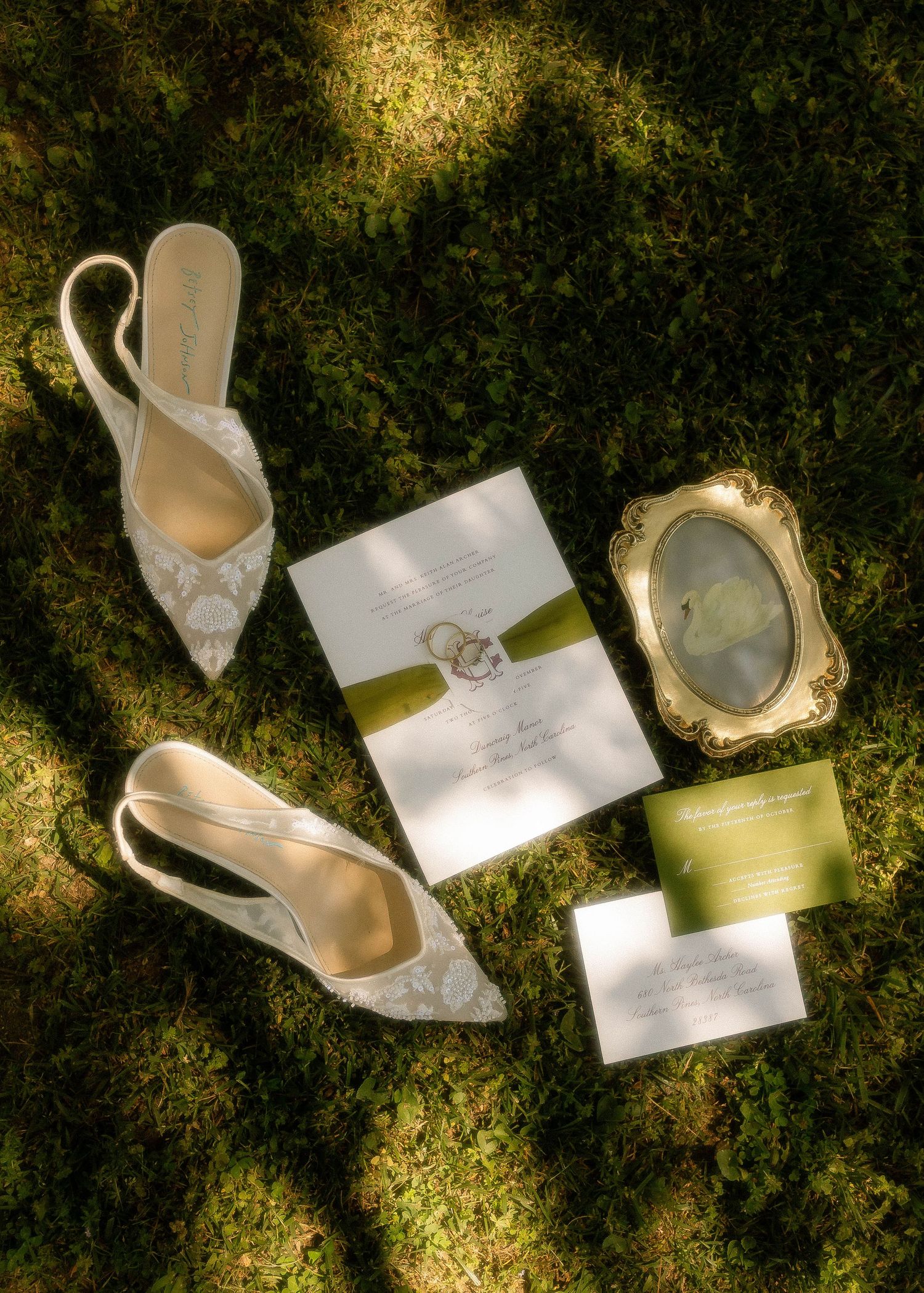 Elegant wedding flat lay on grass featuring lace bridal heels, green ribbon invitation suite, and a gold ornate frame.