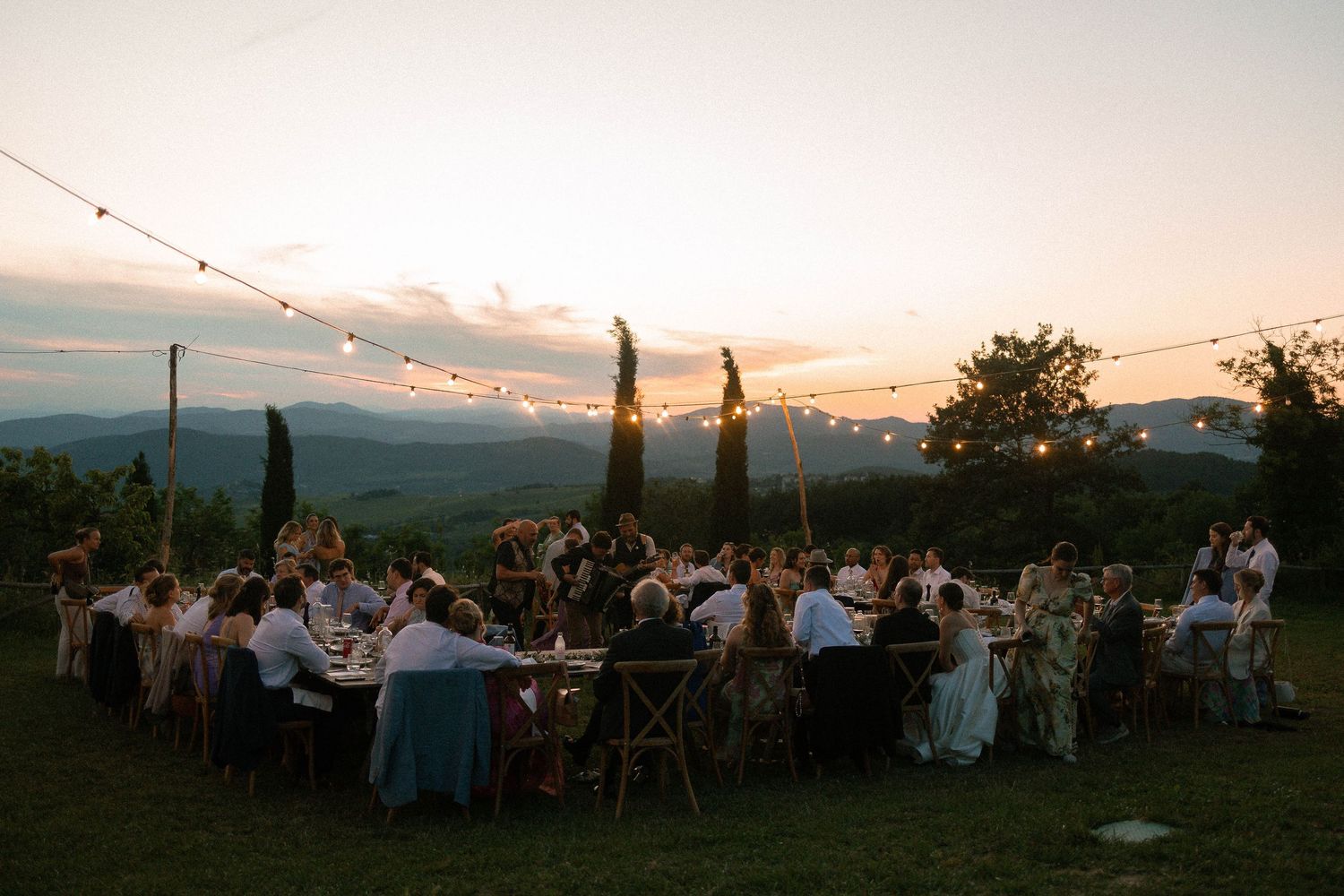 Outdoor wedding reception at sunset with string lights, long dining tables, and scenic mountain views in Tuscany.