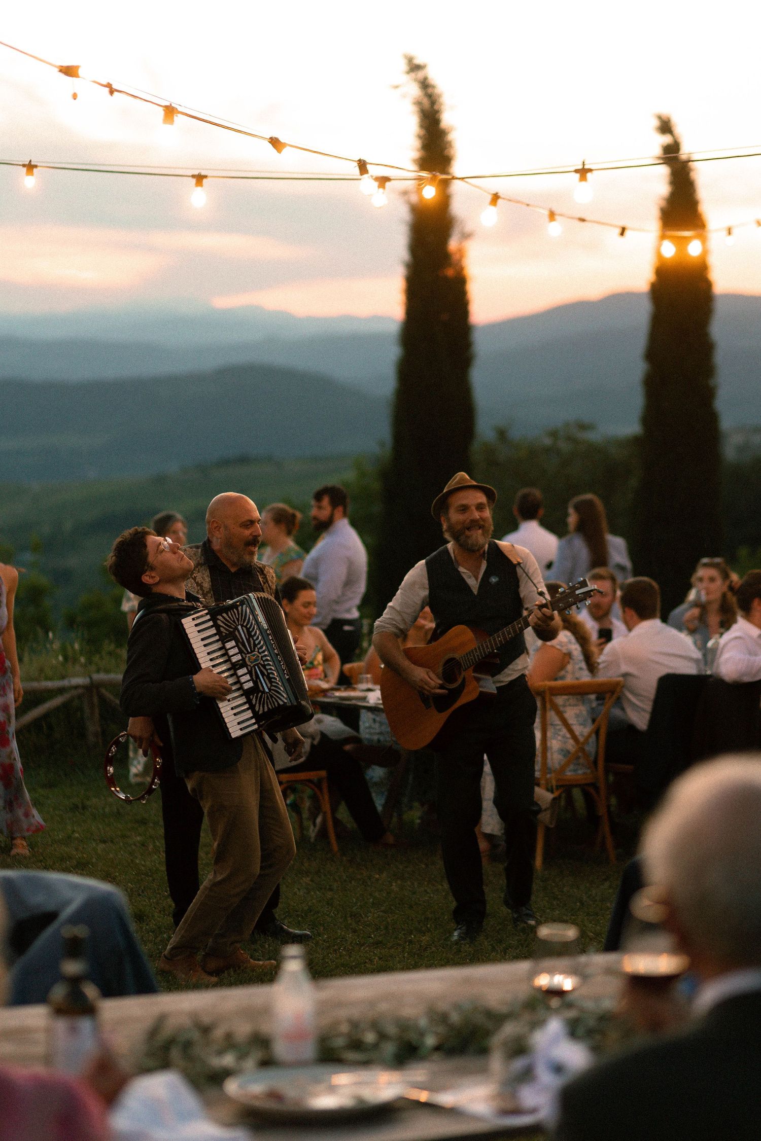 Musicians perform with string instruments and accordion at an outdoor sunset gathering in the Tuscan countryside.