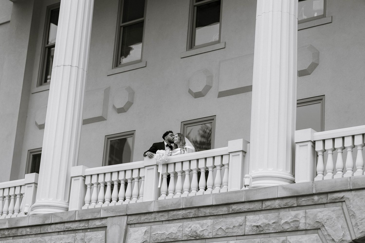 Bride and groom share a romantic moment on a grand white balcony with tall columns of an elegant building.