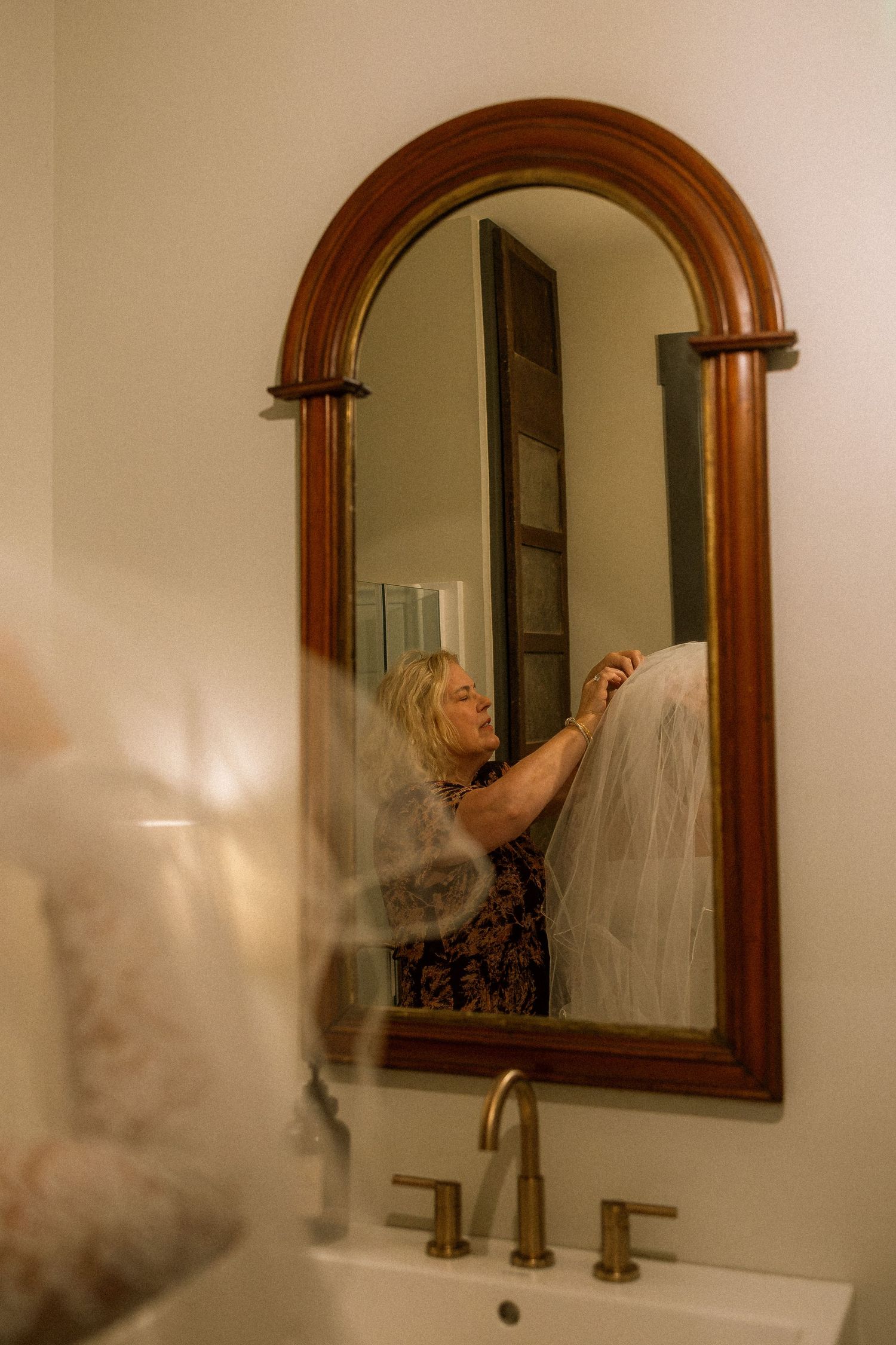 Woman adjusting a bridal veil reflected in an arched wooden mirror above a gold bathroom faucet.