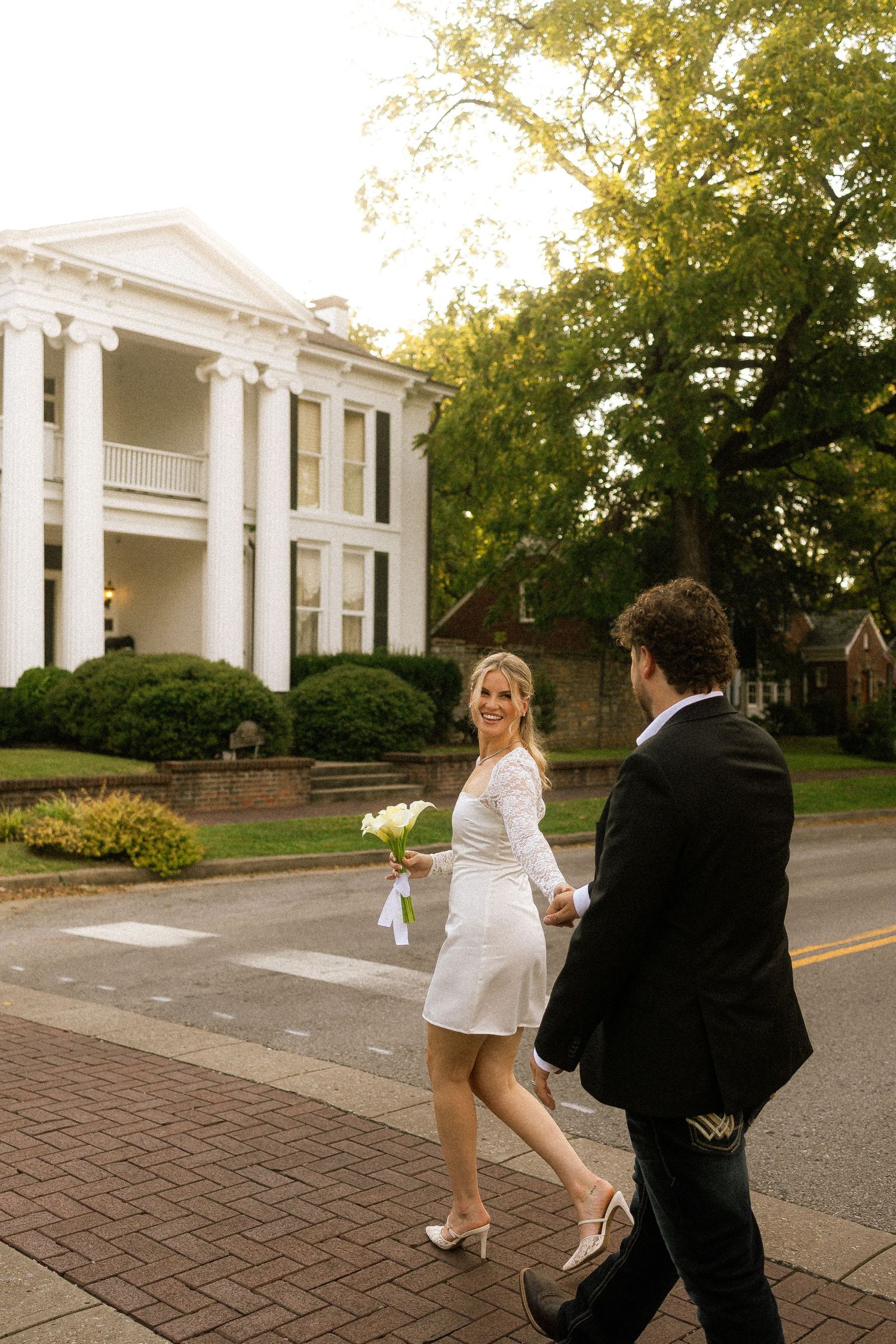Bride in white mini dress holds bouquet while walking with groom in black suit near elegant white columned mansion.