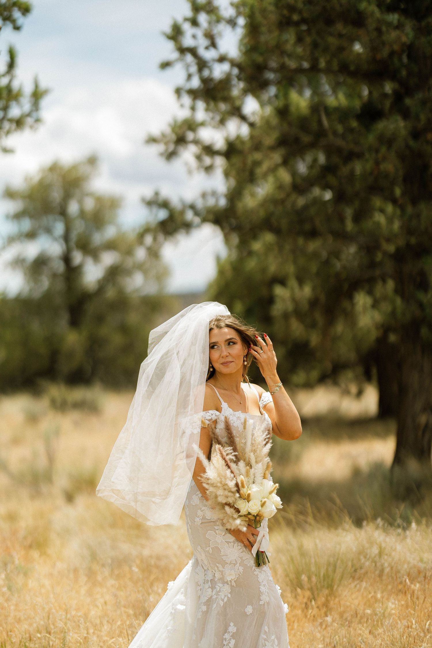 Bride in lace mermaid gown and veil holds boho bouquet in golden field with evergreen trees.