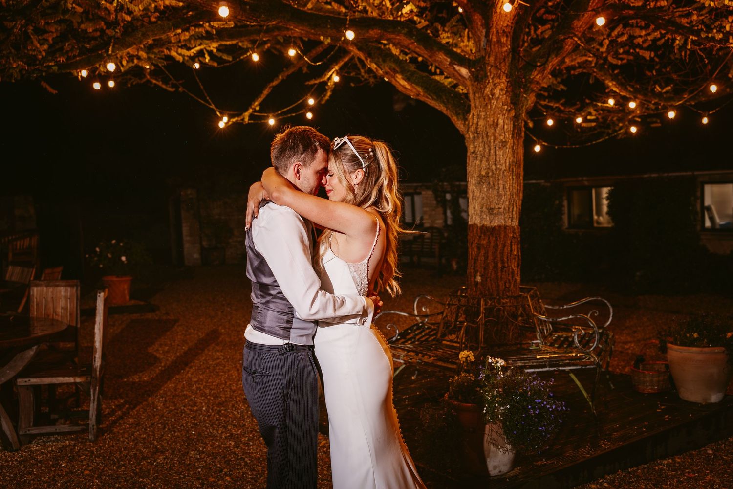 Romantic couple embraces under twinkling lights in a cozy outdoor garden setting at night.