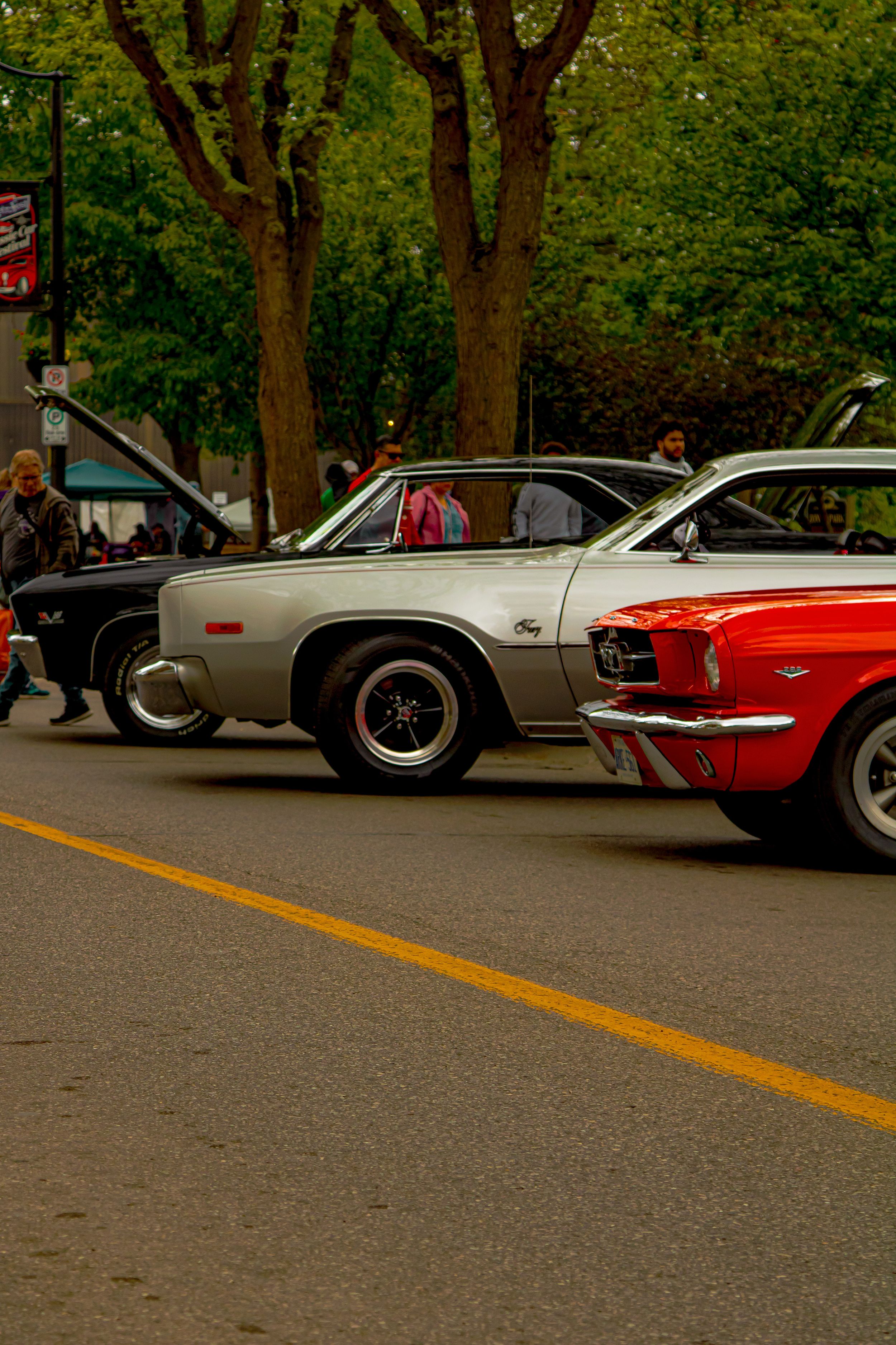 Classic cars including a vintage Plymouth and red Mustang parked on a tree-lined street during a car show.