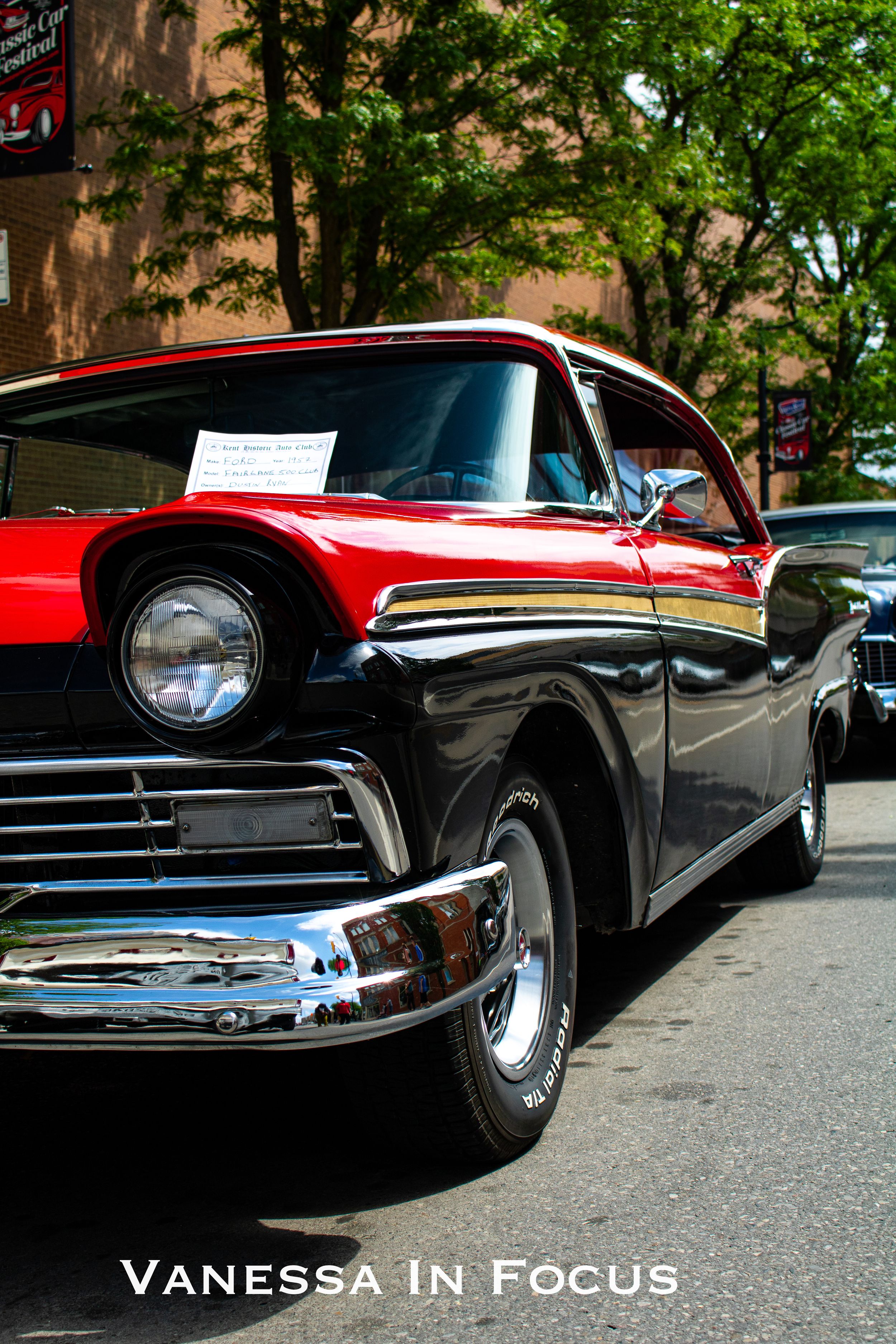 Classic red and black vintage Ford car with chrome trim and headlights parked on a city street with trees in background.