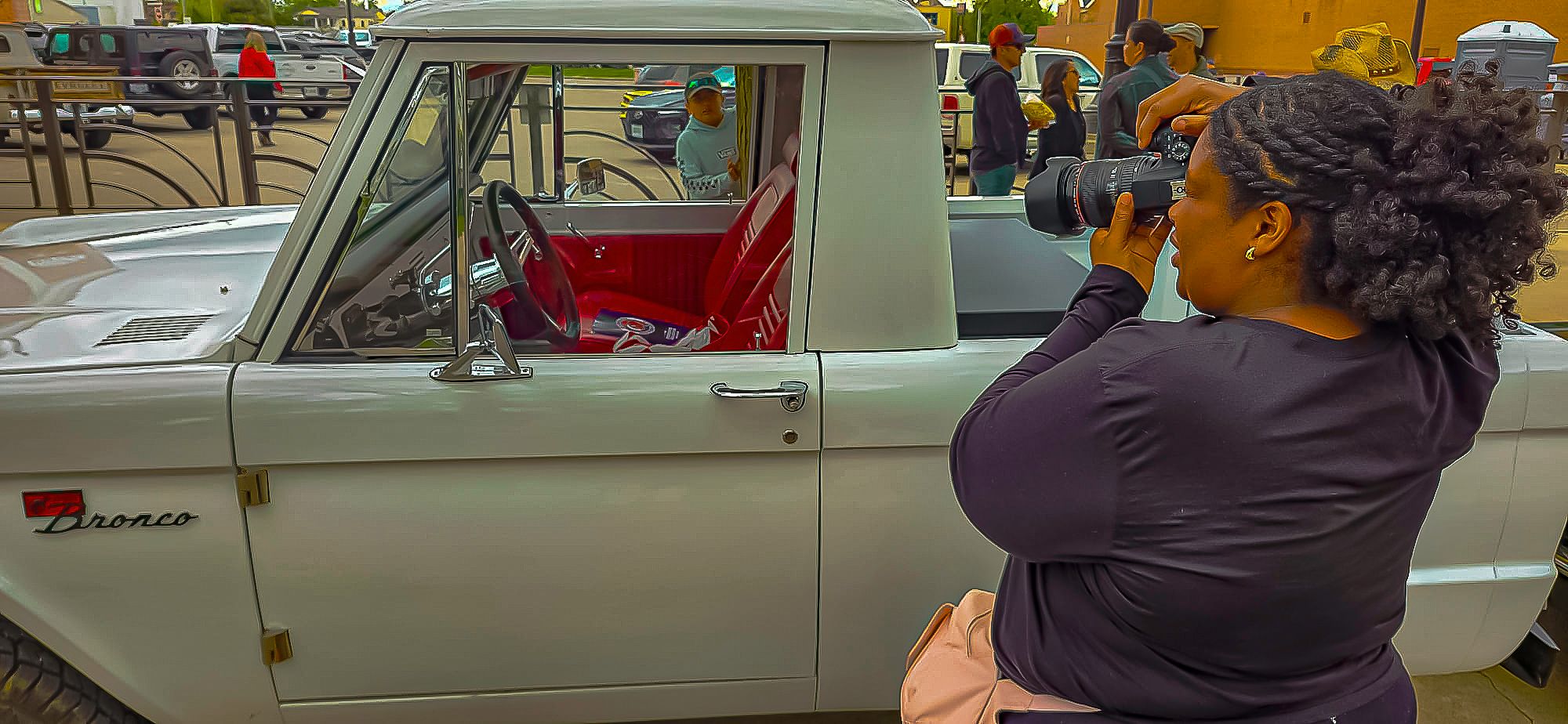 A woman photographs a vintage white Dodge car at an outdoor classic car show on a sunny day.