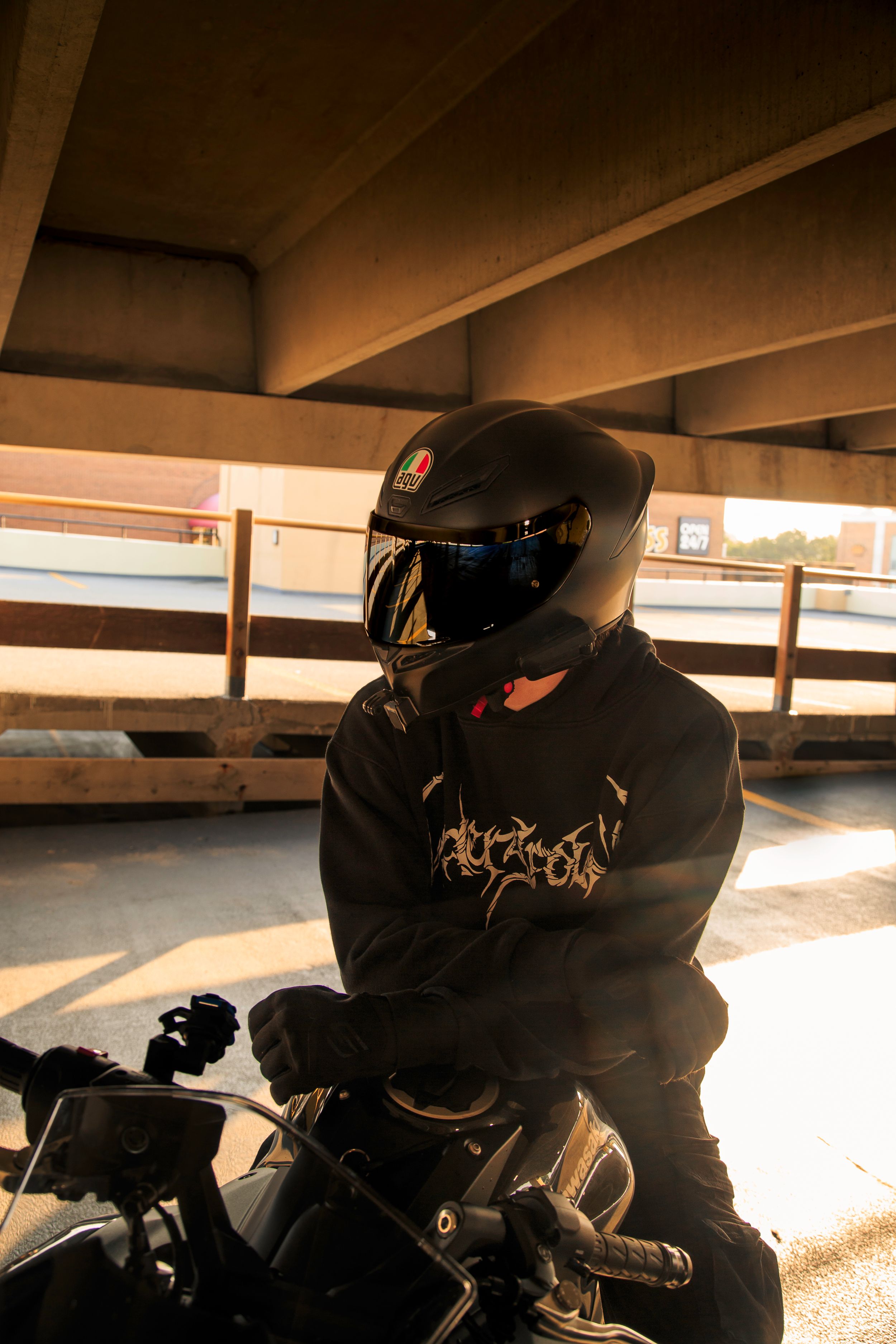 Motorcyclist in a black helmet and graphic hoodie sits on a motorcycle under a concrete overpass in warm golden sunset light.