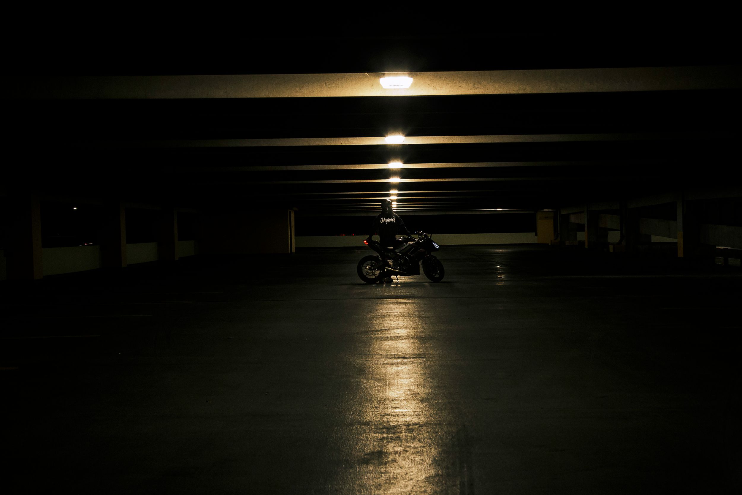 Lone motorcycle parked in a dark, empty parking garage with overhead lights casting a glowing reflection on the concrete floor.