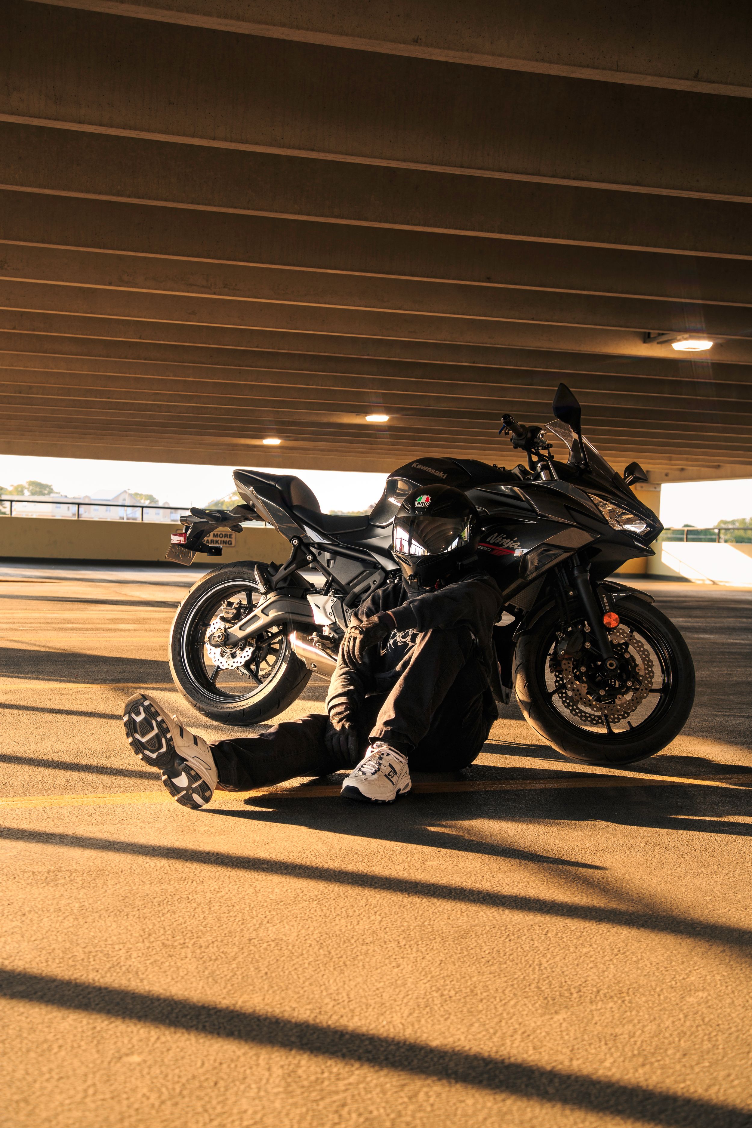 Rider in black gear and helmet sits on the ground leaning against a black sport motorcycle under a parking garage at golden hour.