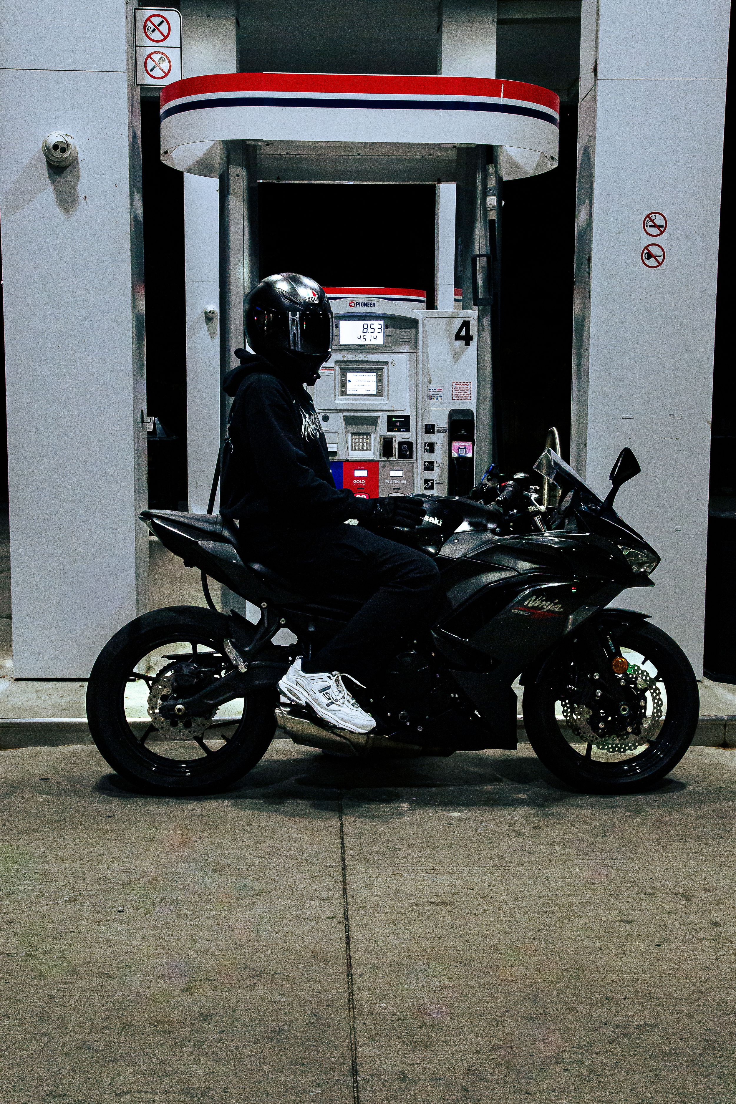 Rider in a black helmet and hoodie sits on a black sport motorcycle beside a lit gas station pump at night.