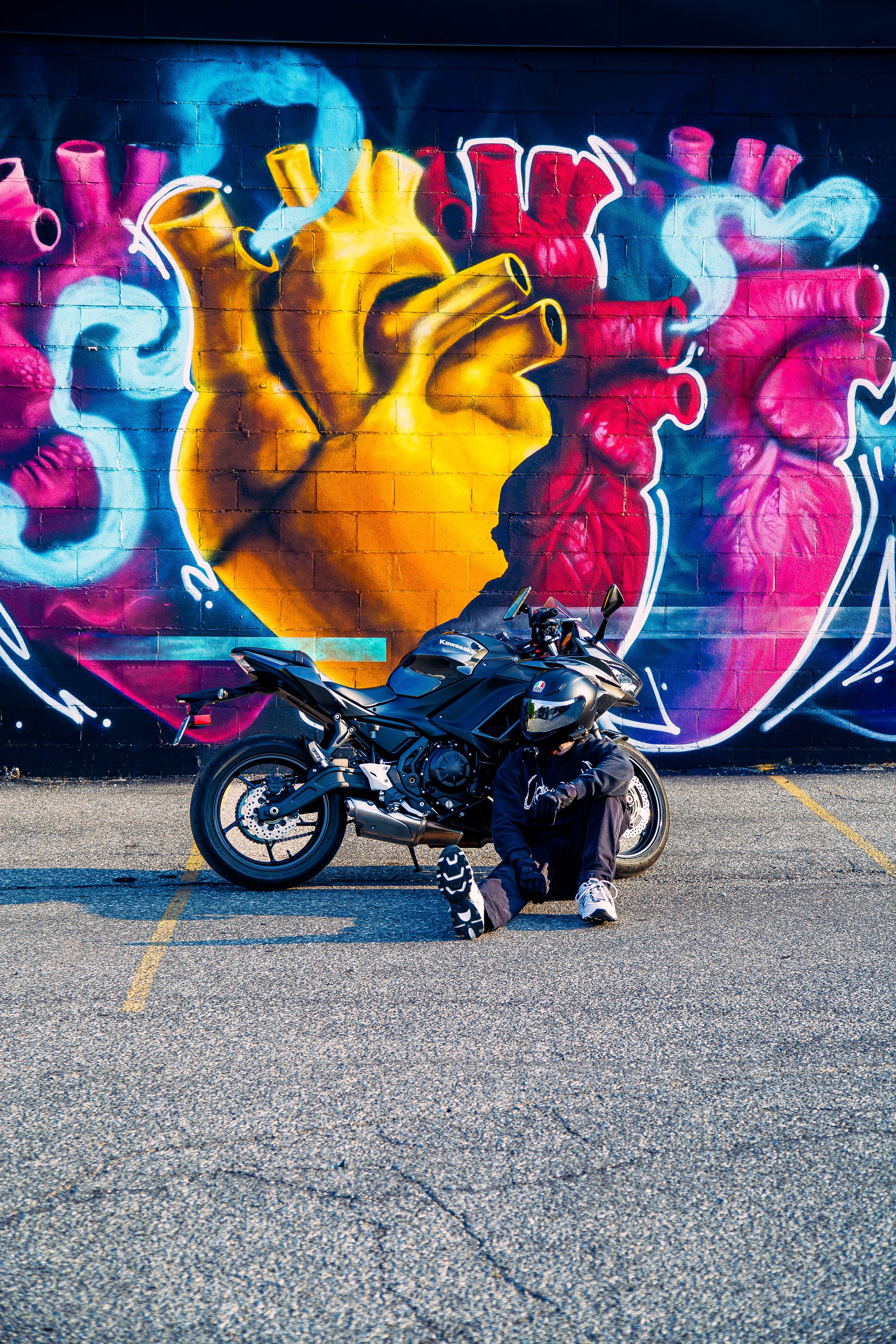 Rider crouches beside a black sport motorcycle parked in front of a colorful graffiti mural of glowing anatomical hearts on a brick wall.