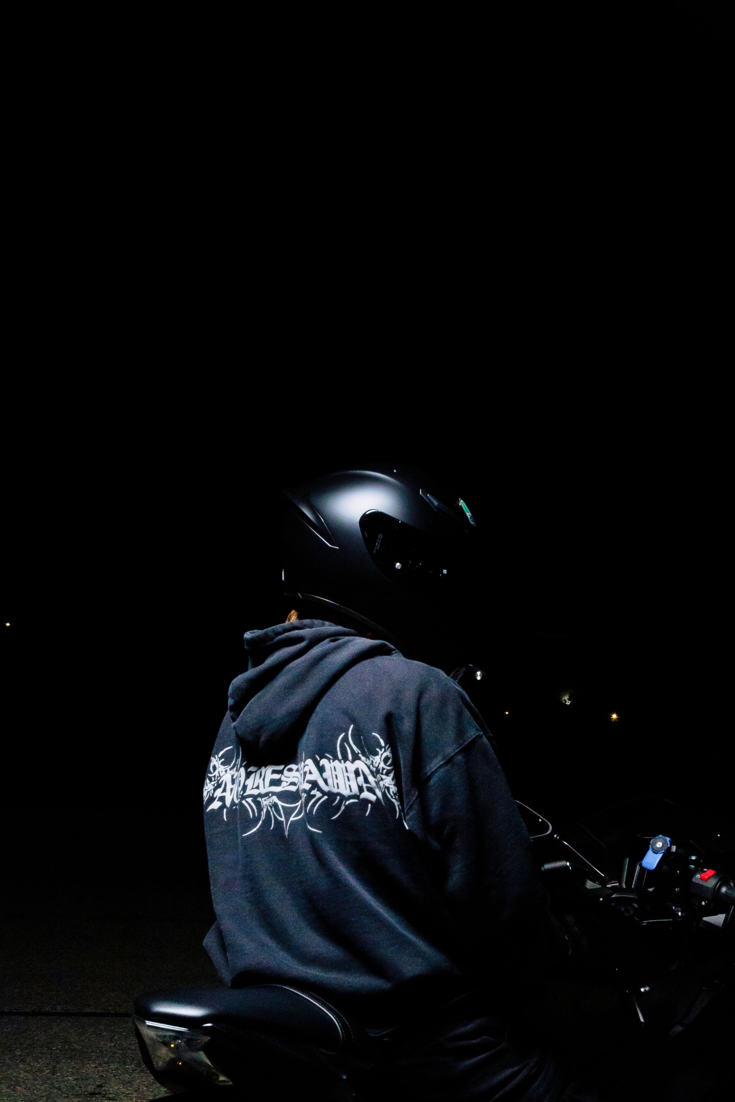 Motorcyclist in a black helmet and gray hooded sweatshirt with graphic lettering sits on a motorcycle against a dark night background.