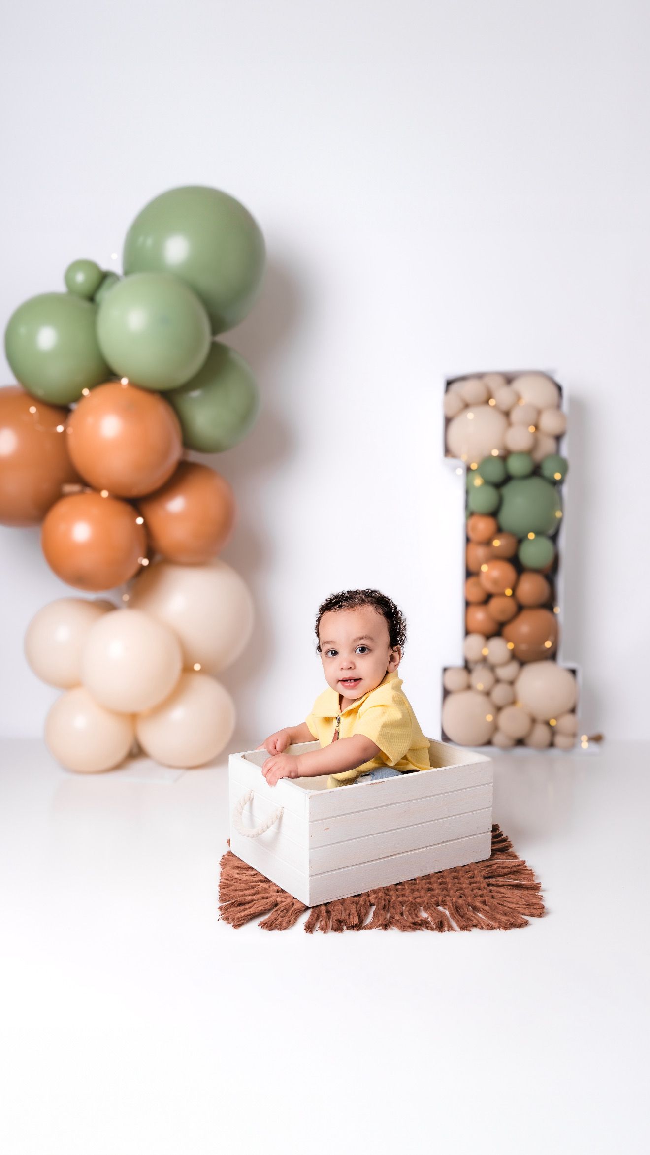 Baby in yellow outfit posed in white box with green and peach balloon decorations.