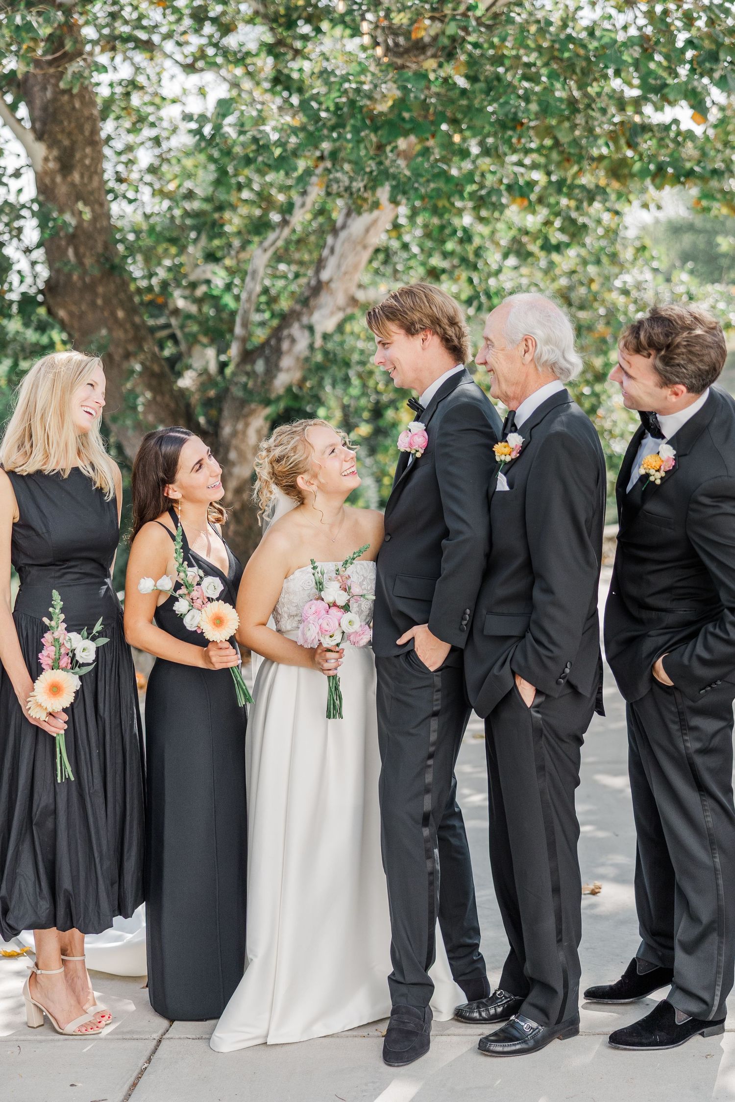 Wedding party members in formal black attire interacting and laughing together outdoors.