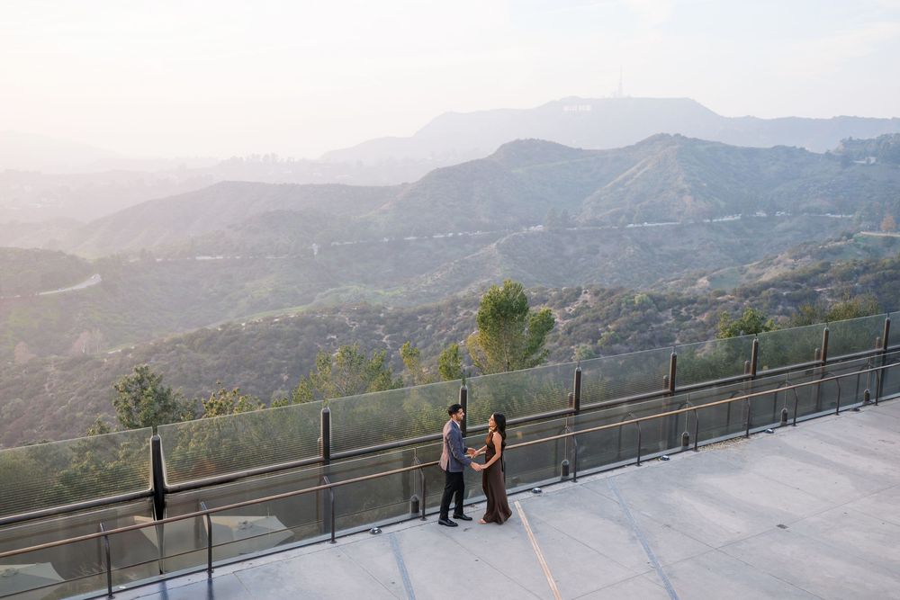 Two people stand at a scenic mountain overlook with metal railings during sunset in a hazy mountainous landscape.