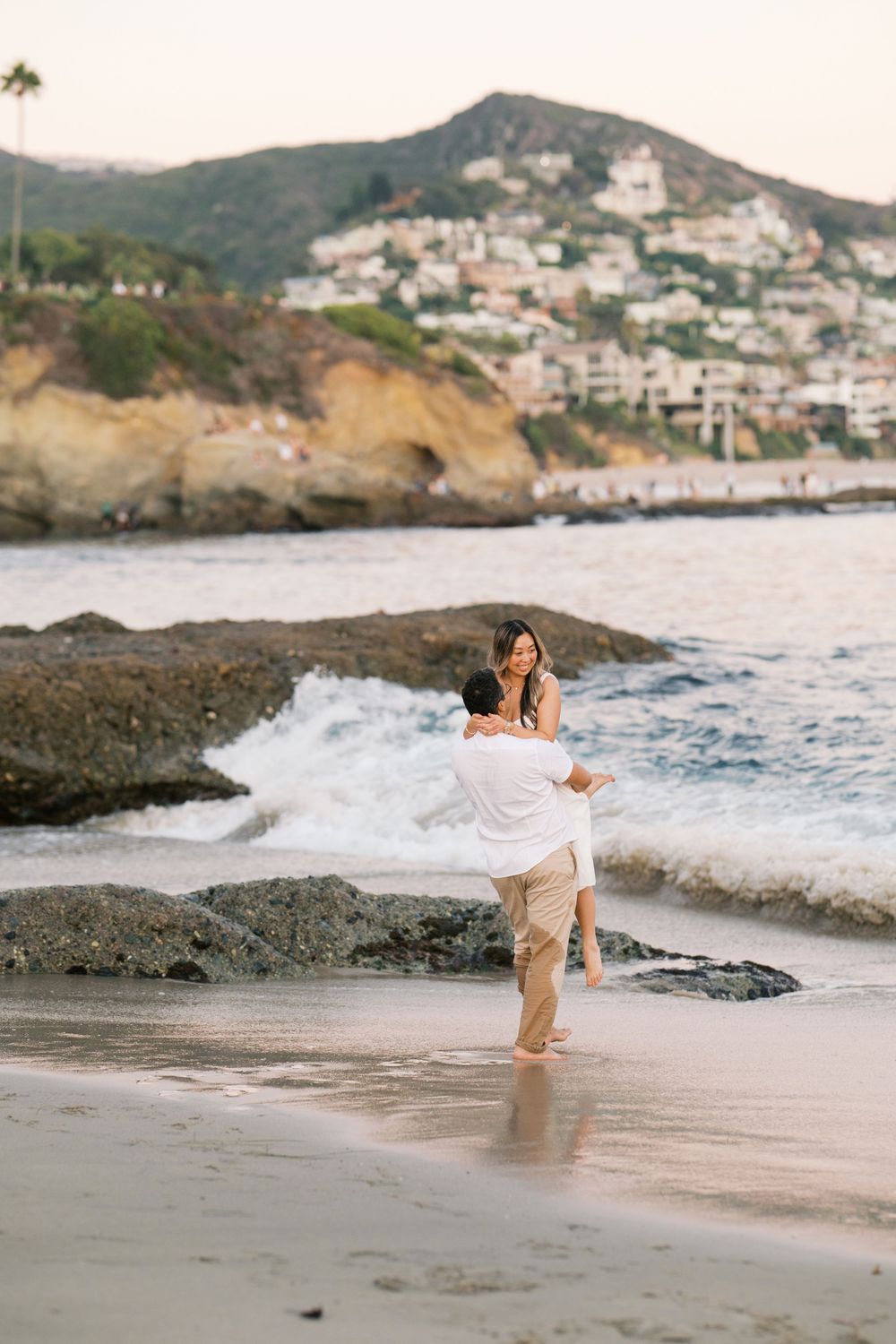 Couple embraces on rocky beach at sunset with coastal homes on hillside in background.