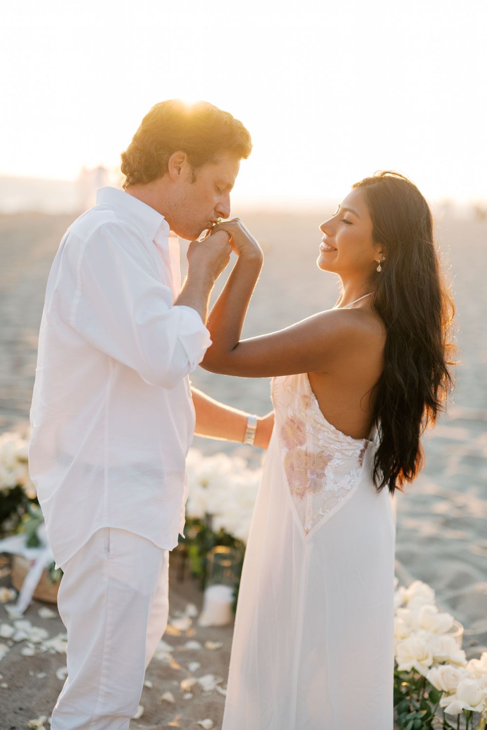 Romantic couple in white outfits dancing together on a sunny beach at sunset.